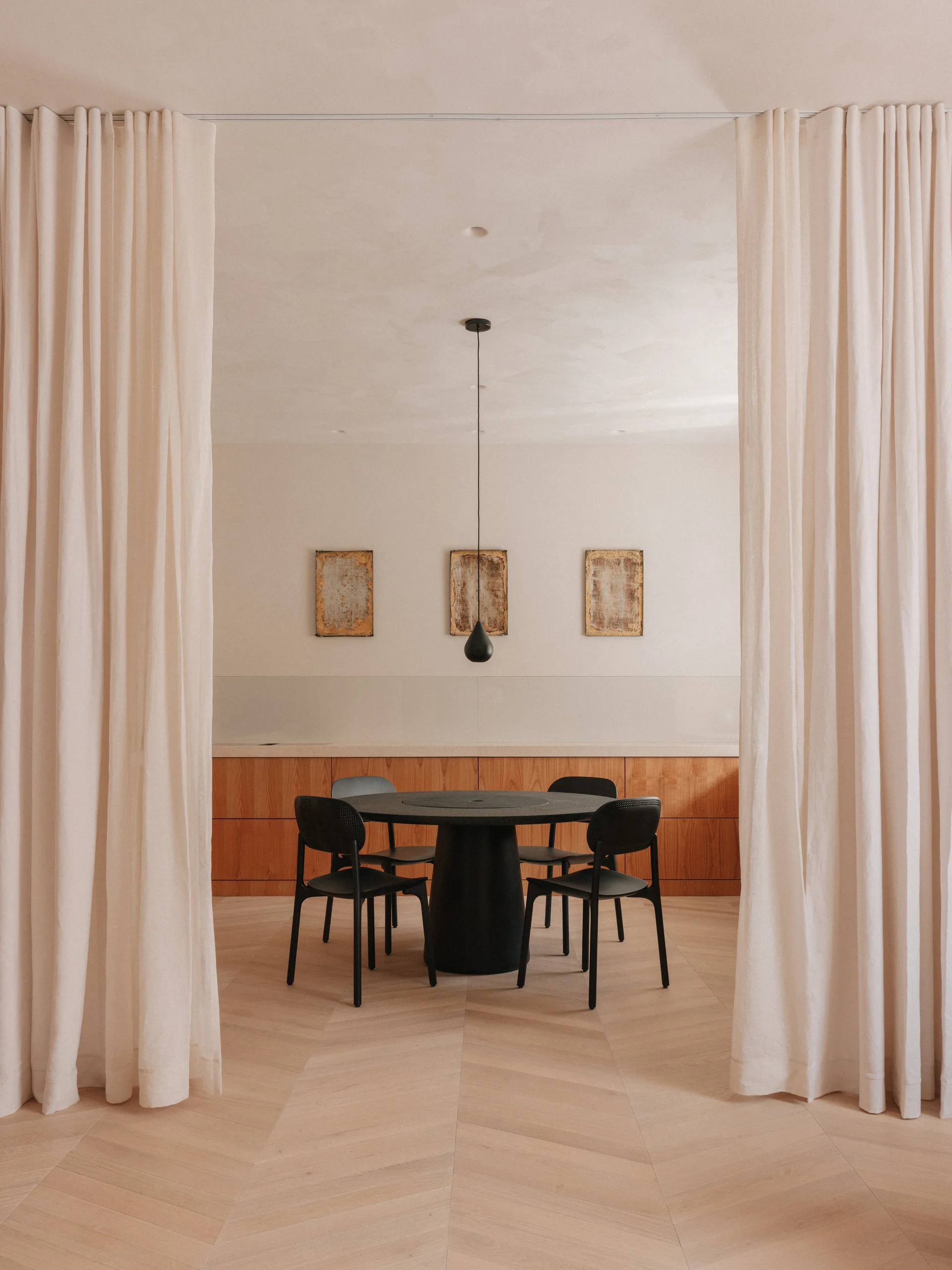 A modern dining room featuring a black round table and chairs framed by light beige curtains on a herringbone wood floor.