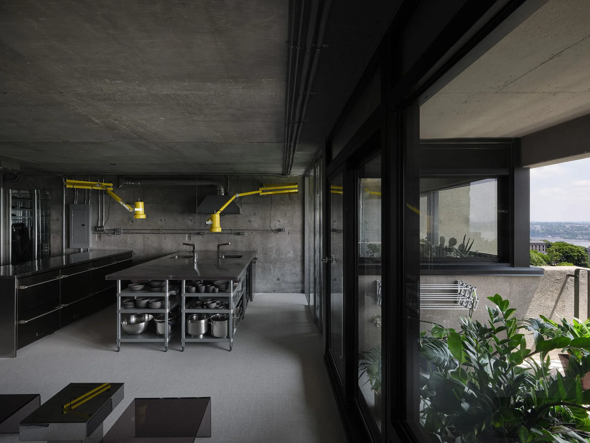 An industrial kitchen features raw concrete walls, a steel island with open storage, black cabinets, and bright yellow articulating wall lamps.