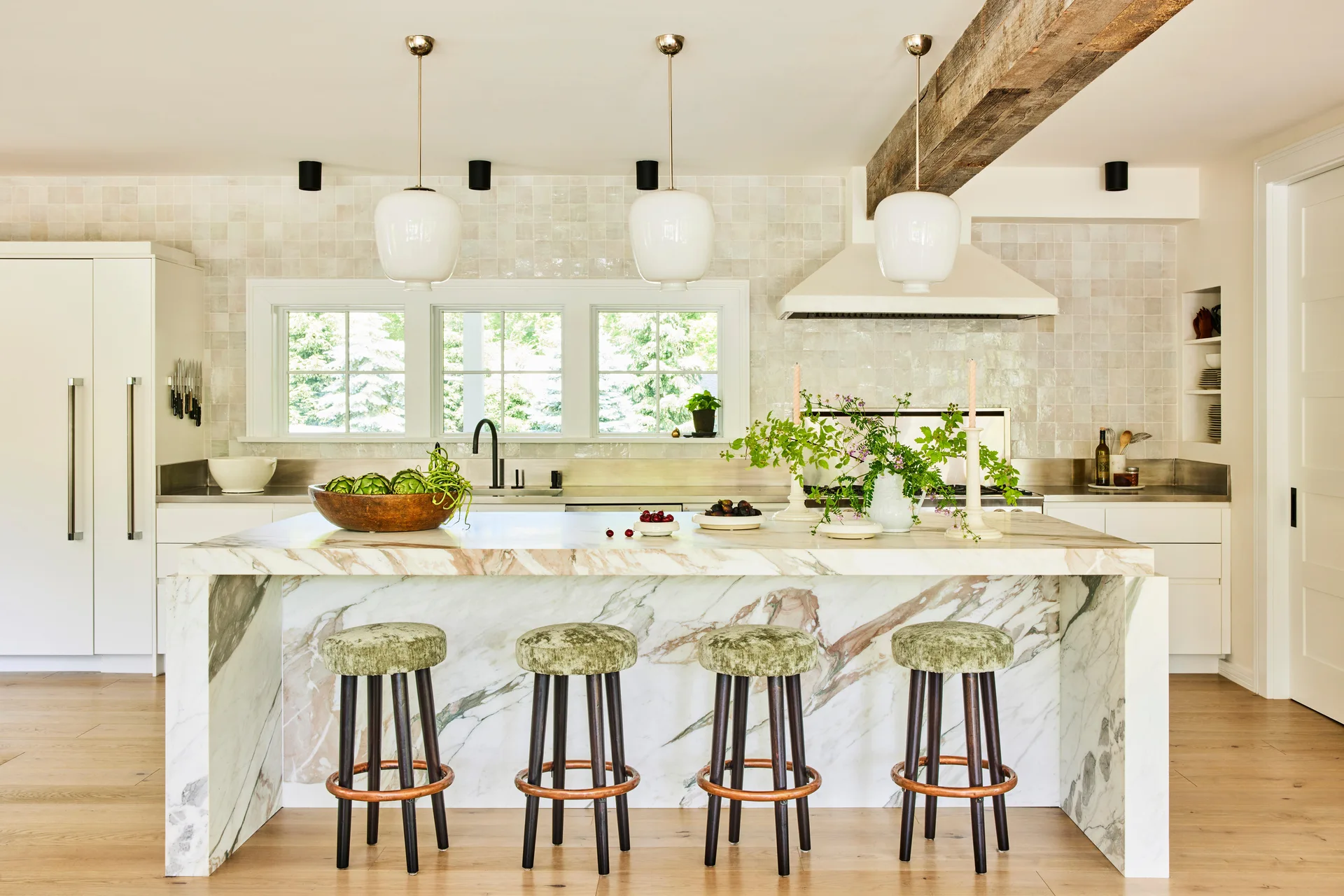 A modern kitchen featuring a marble waterfall island with green velvet bar stools under white pendant lights and an exposed wood ceiling beam.
