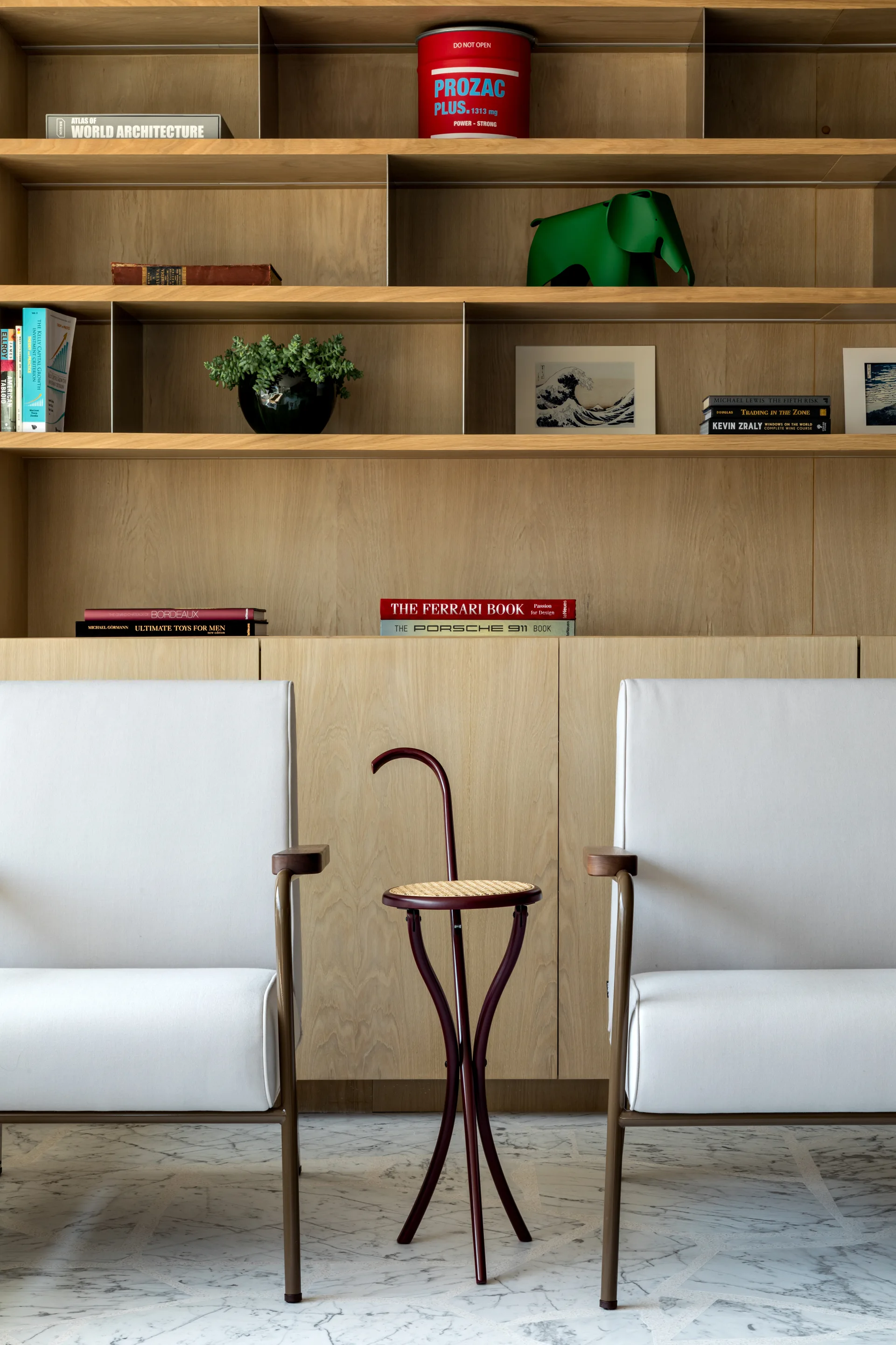 Two white modern armchairs flank a sculptural dark wood side table with a cane seat in front of a light oak built-in bookcase on marble flooring.