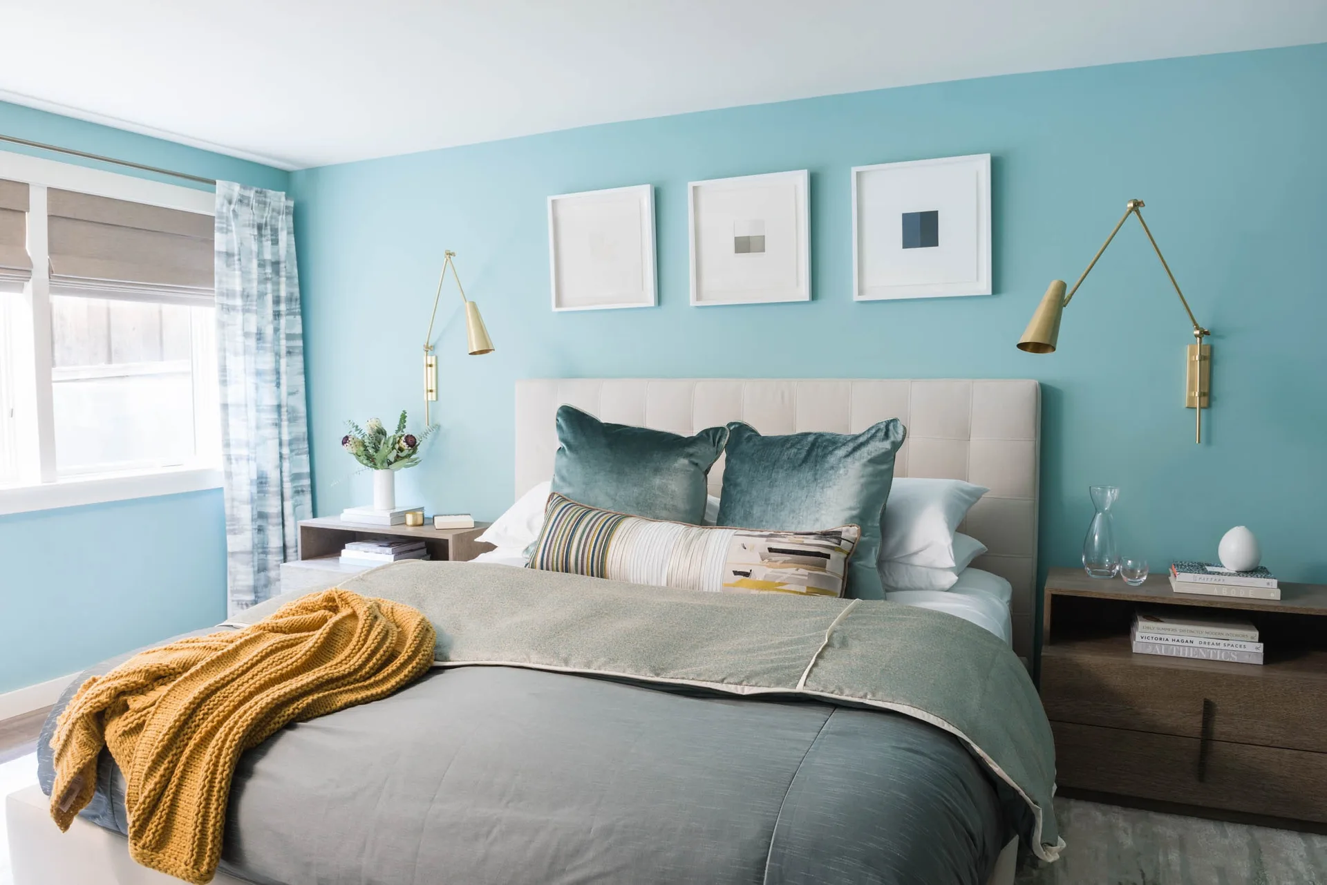 A bedroom with light blue walls features a beige tufted headboard, brass swing-arm sconces, and layered blue bedding with a mustard yellow throw.