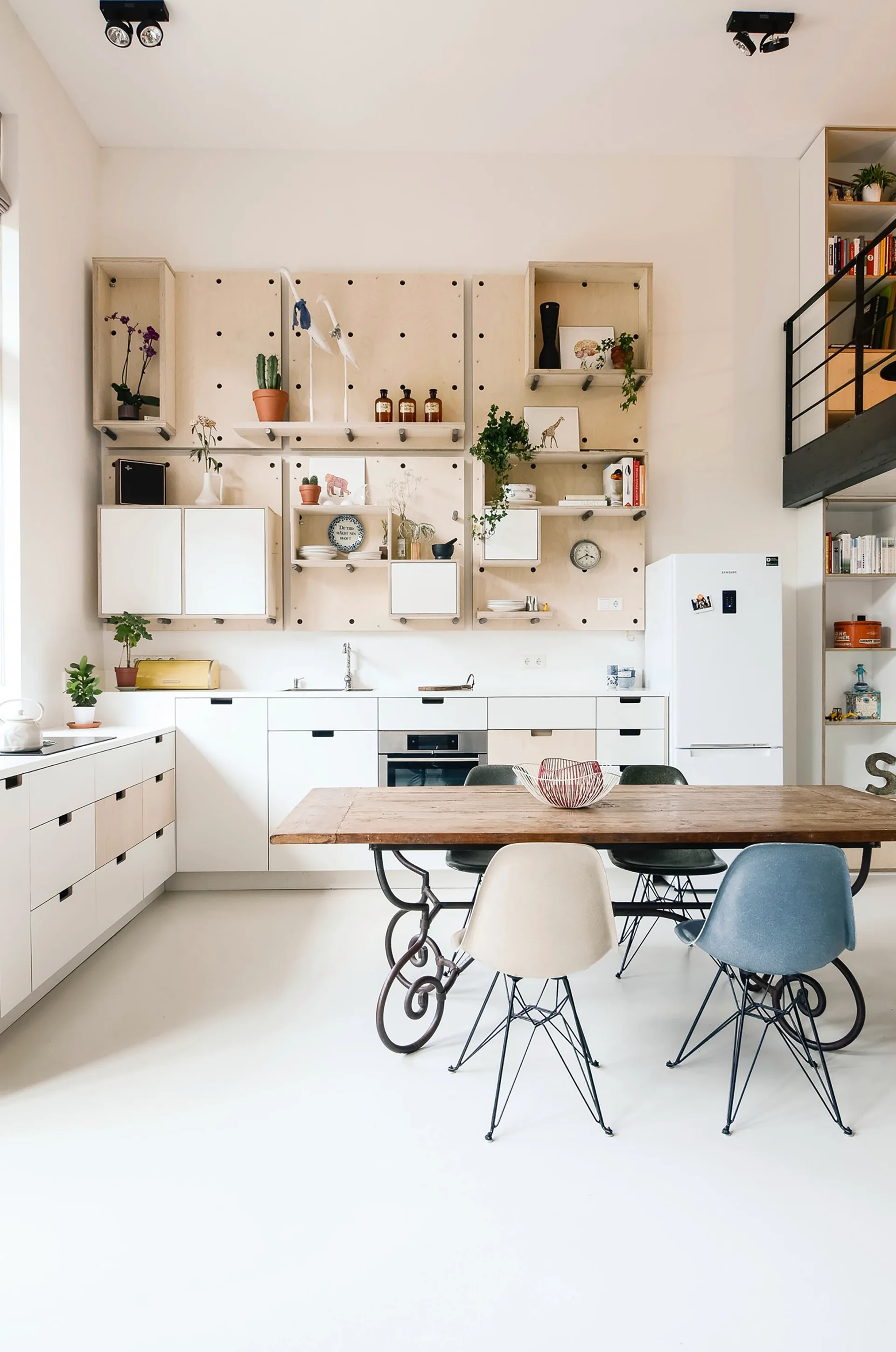 A bright kitchen features white lower cabinets, a plywood pegboard shelving wall, and a rustic wooden dining table with molded chairs.