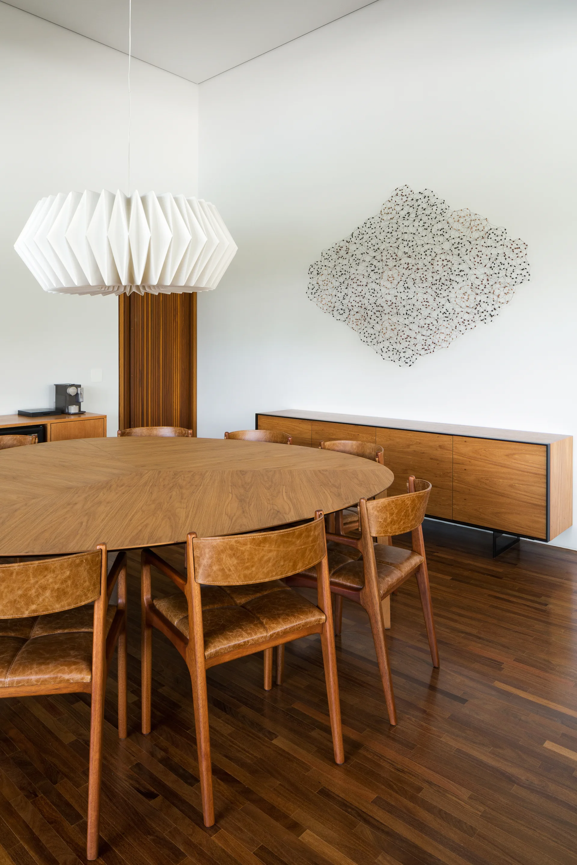 A round wooden dining table surrounded by leather chairs sits under a white pleated pendant light next to a wooden sideboard with wire wall art.