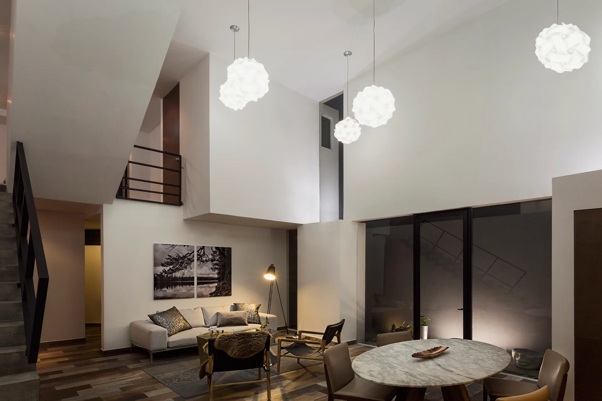 A modern double-height living room featuring a light grey sofa, round marble dining table, and three white sculptural pendant lights.