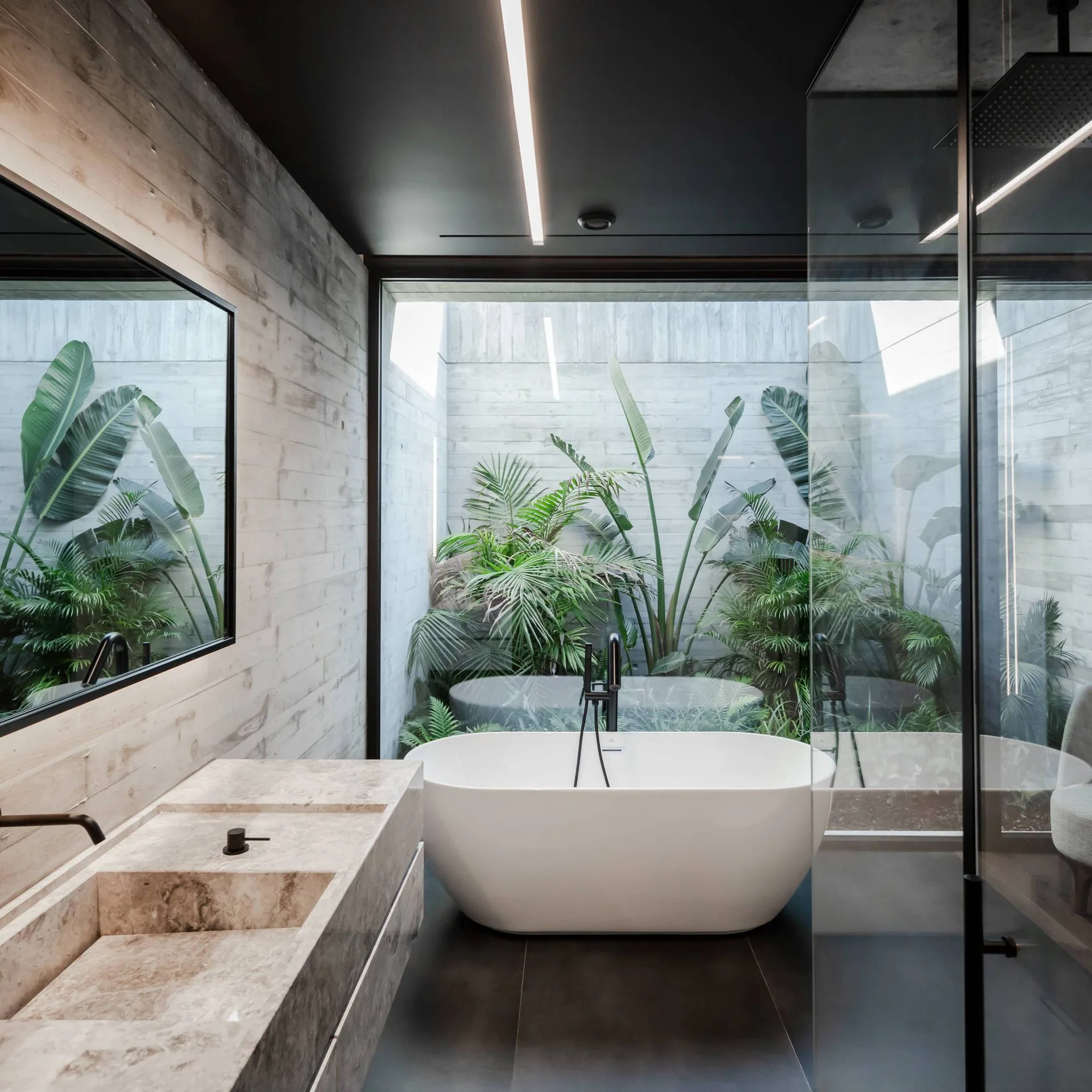 A modern bathroom features a stone vanity, a freestanding white bathtub, and a large glass window looking out onto lush green tropical plants.