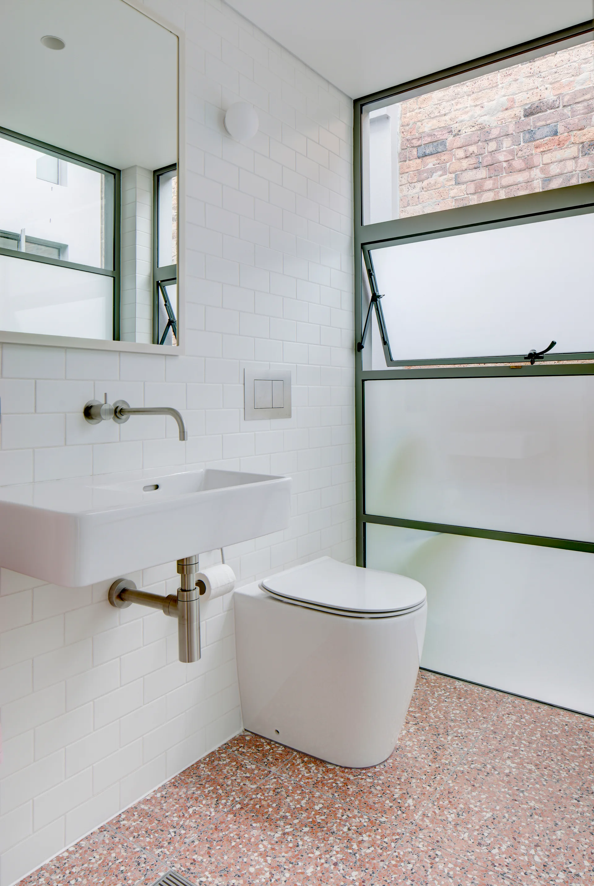 A modern bathroom with white subway tile walls, a wall-mounted ceramic sink, a back-to-wall toilet, and terracotta terrazzo flooring.