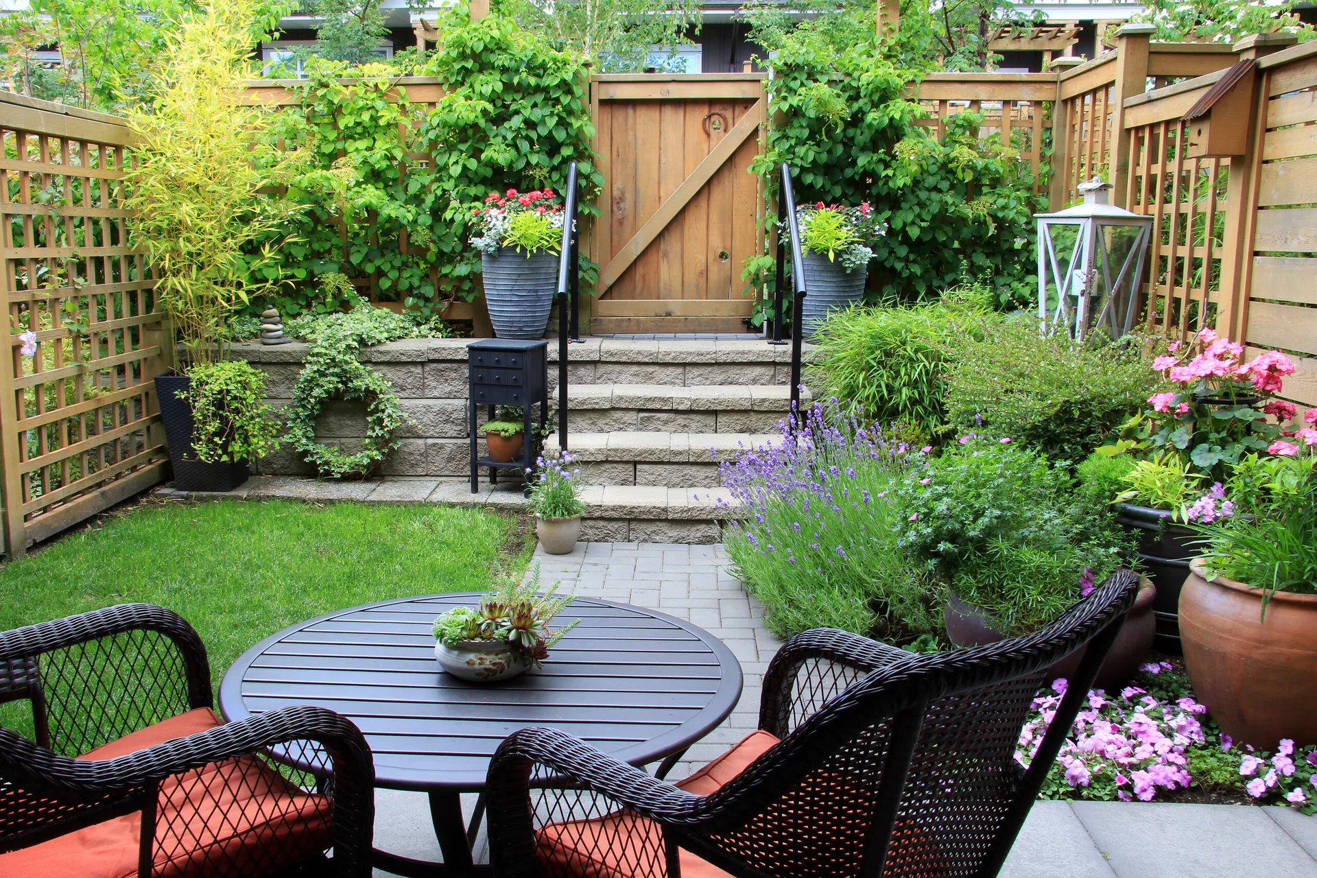 A small backyard patio features a wicker bistro set and stone stairs leading up to a wooden gate nestled among lush garden plantings.