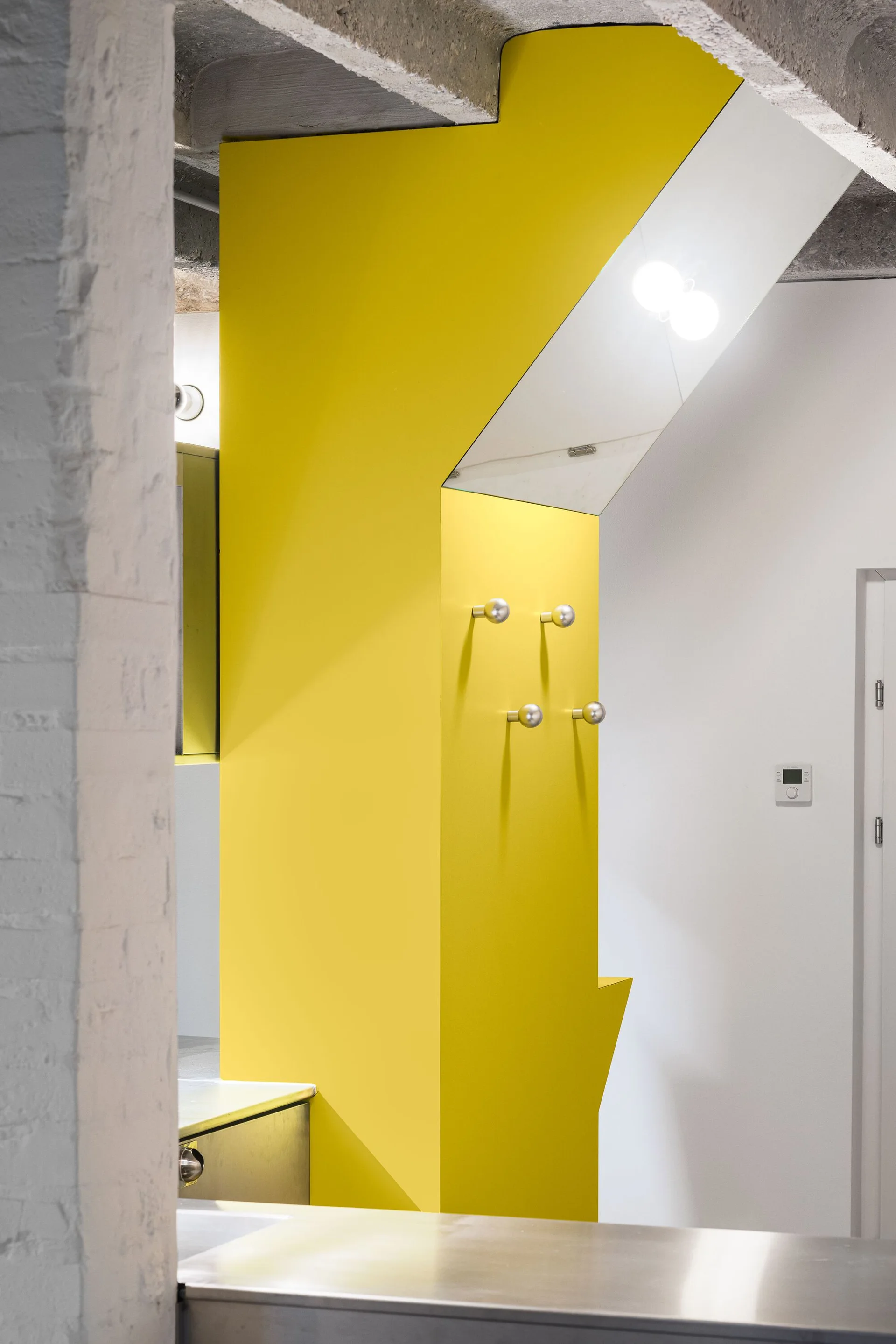 A bright yellow architectural wall panel with four round silver coat hooks stands between a raw concrete ceiling and a white wall.