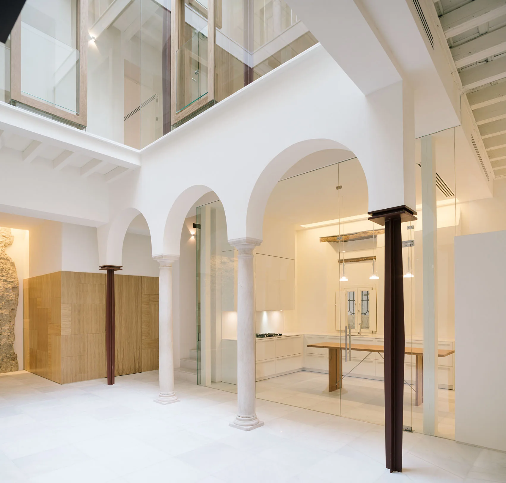A modern white kitchen enclosed by glass partitions sits behind a series of historic white stone arches and columns.