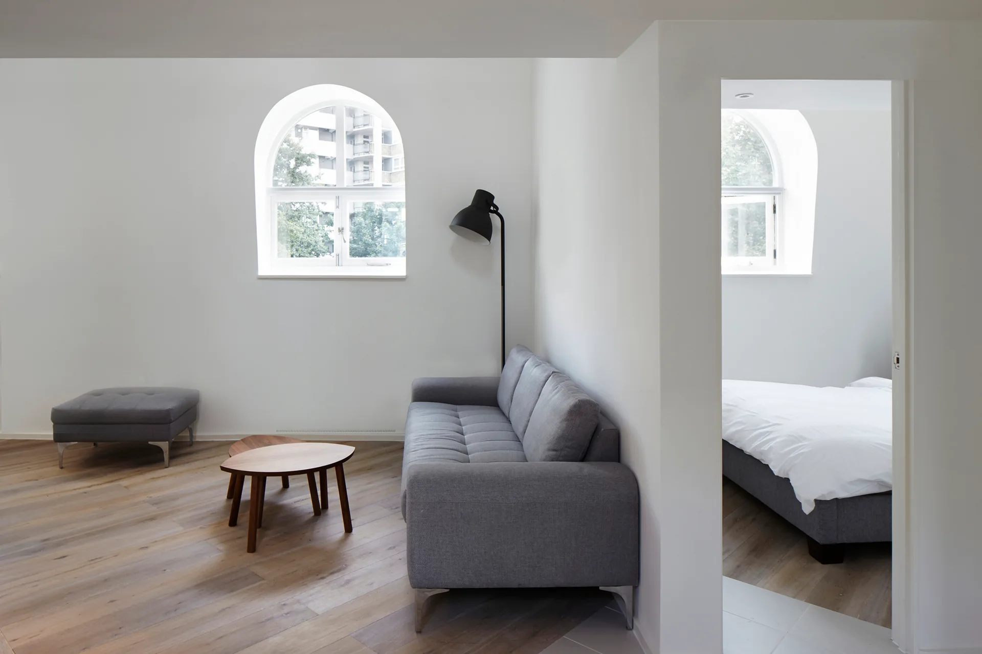 A minimalist white living room features a grey sofa, nesting wooden coffee tables, and an arched window, with a bedroom visible through an open doorway.