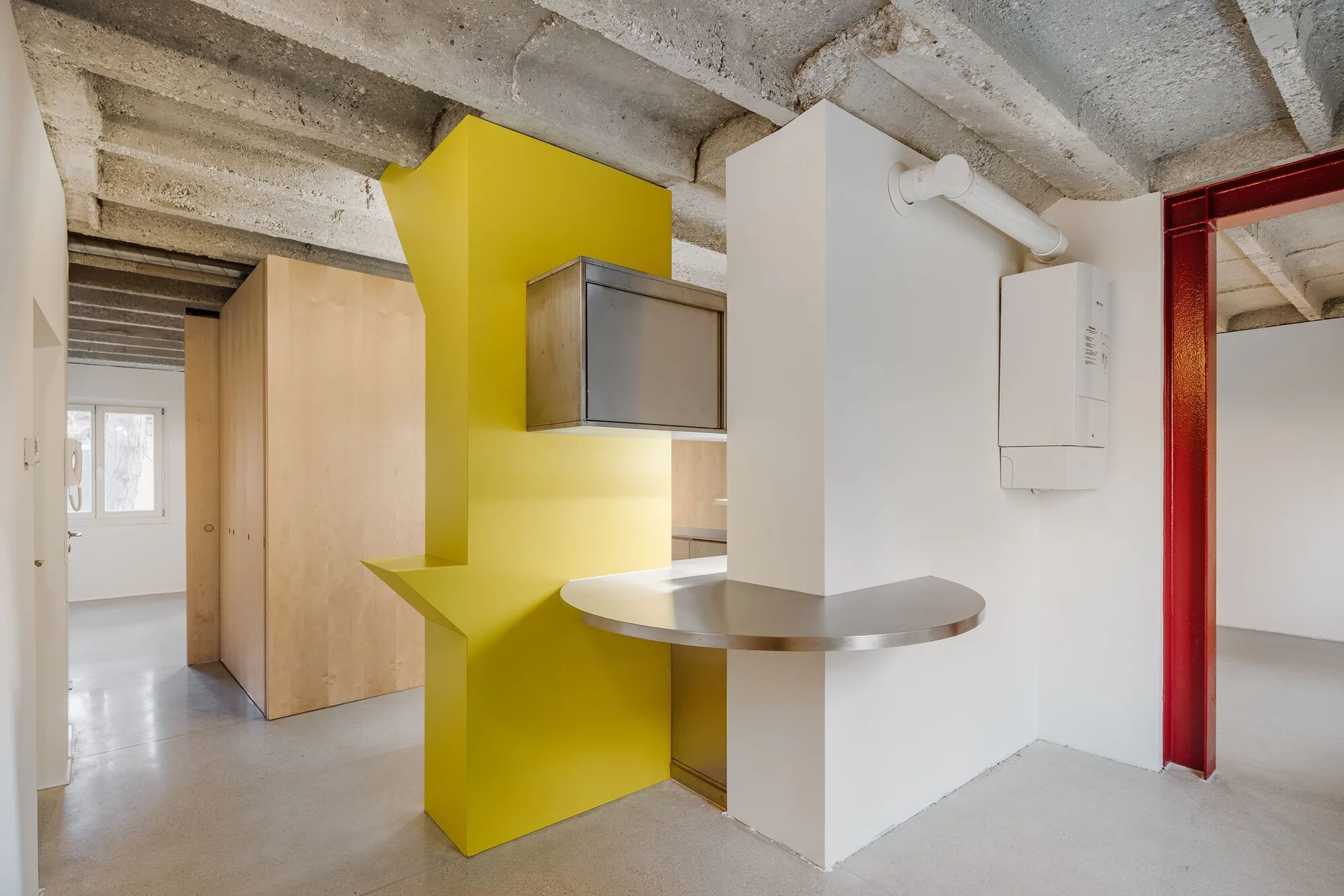 A minimalist kitchen features a custom yellow geometric cabinet structure, a curved stainless steel countertop, and an exposed concrete ceiling.