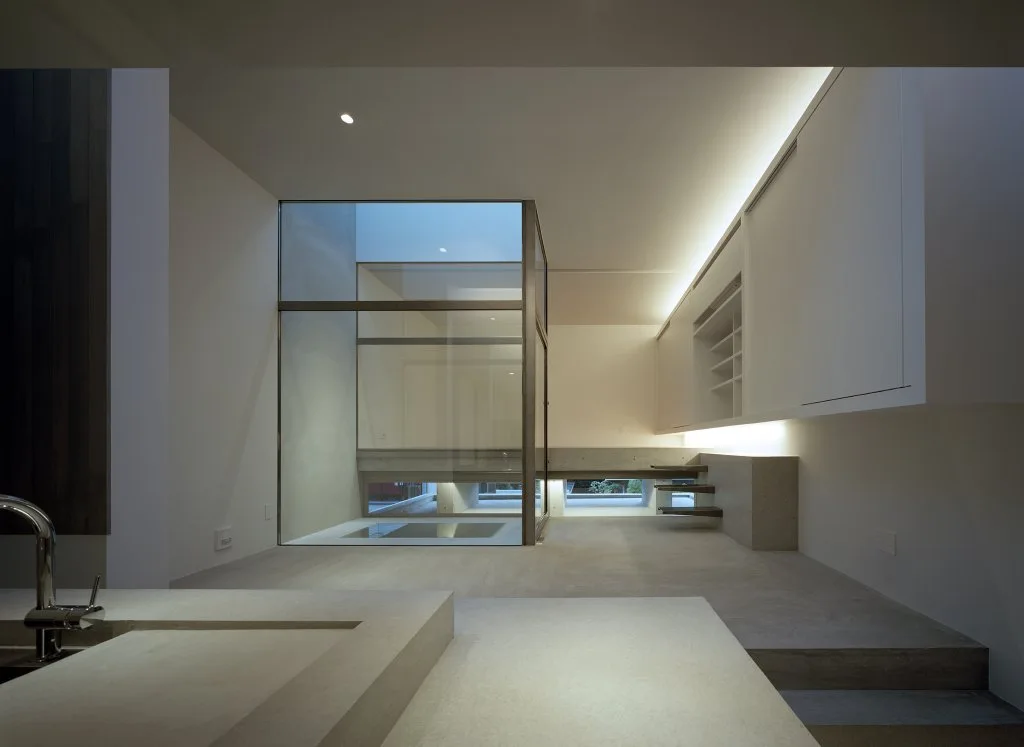 A minimalist grey concrete room featuring a central glass-walled enclosure, built-in white cabinetry, floating wooden stairs, and soft cove lighting.