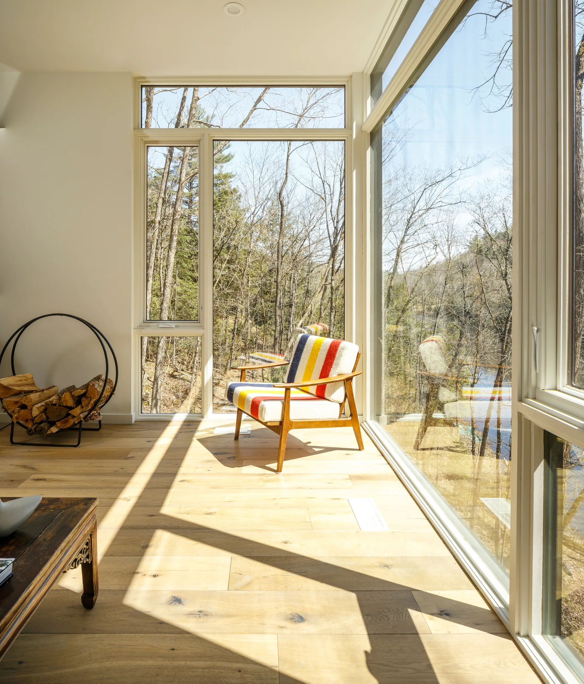 A wooden mid-century armchair with striped upholstery sits on light hardwood floors next to large windows overlooking a forest.
