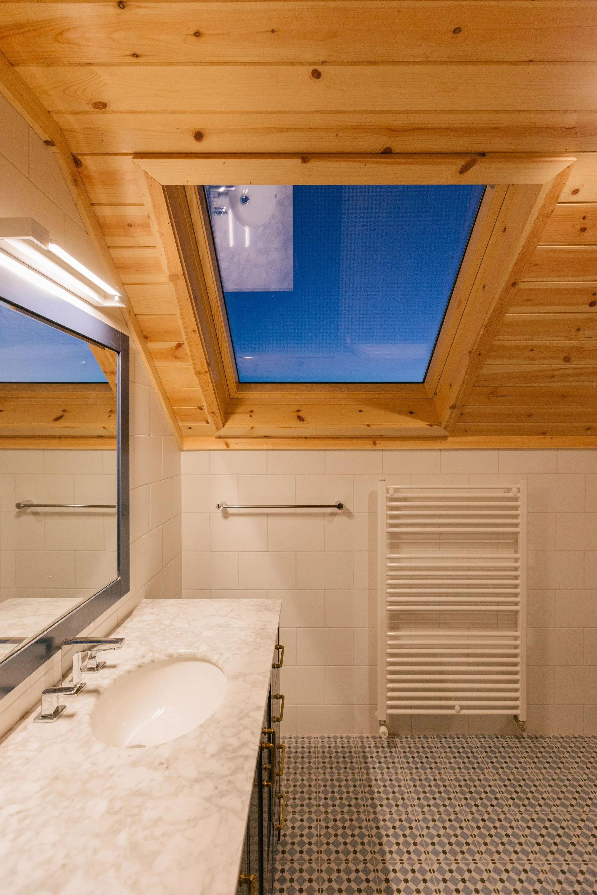 A modern bathroom features a sloped wooden ceiling with a skylight, a marble vanity, and blue and white patterned floor tiles.