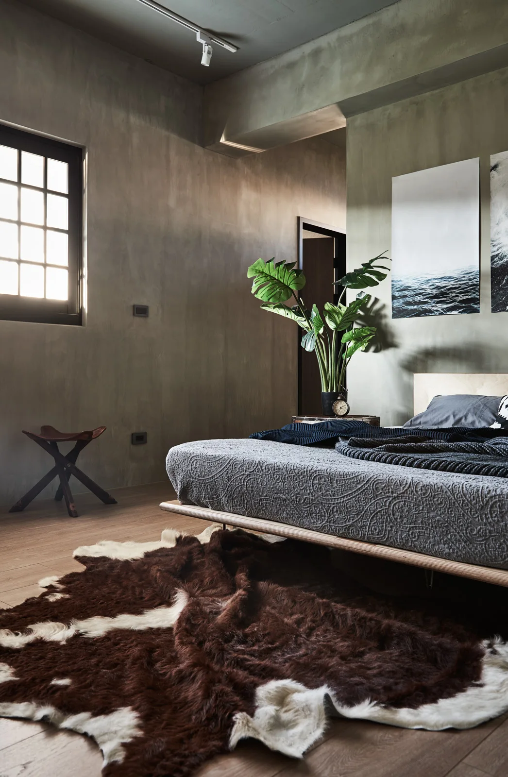 A modern bedroom features a low wooden platform bed on a brown cowhide rug against textured olive green walls next to a grid window.