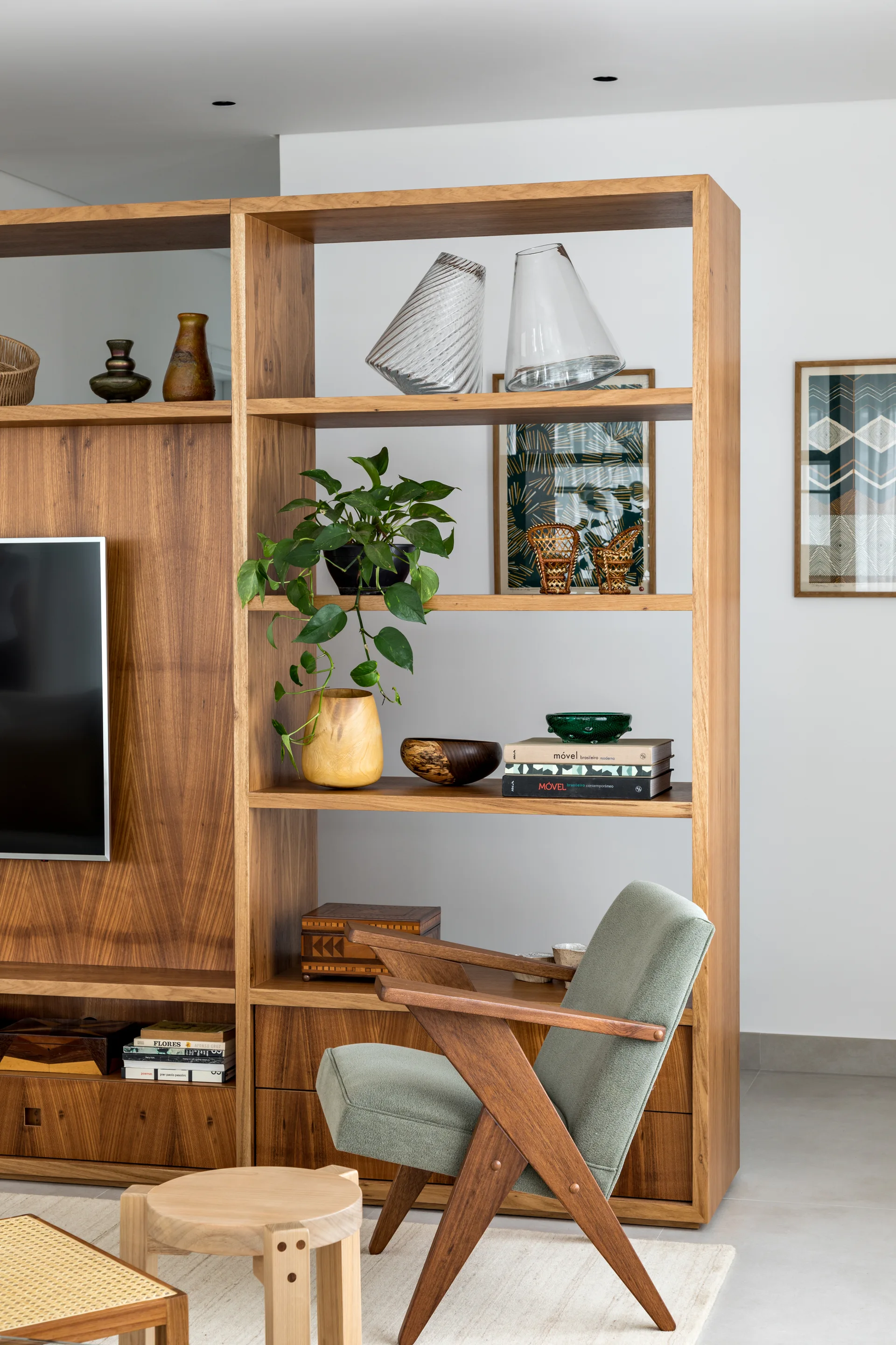 A warm wood open shelving unit decorated with books and a pothos plant stands next to a sage green mid-century modern armchair.