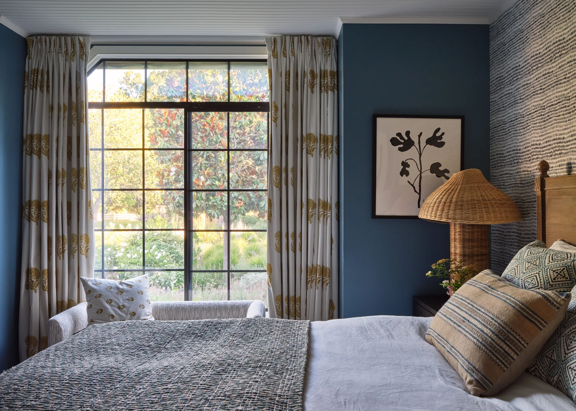 A bedroom corner features a bed with patterned pillows, a rattan table lamp against a blue wall, and a large window with patterned drapes.