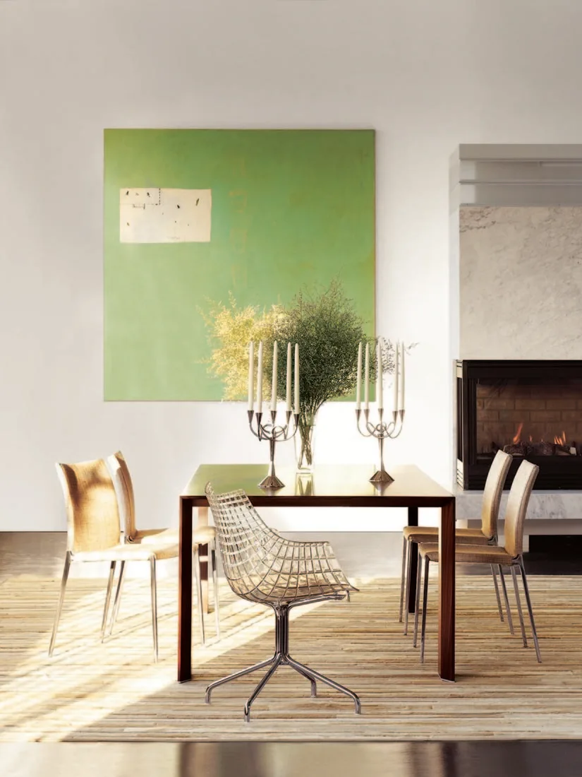 A contemporary dining room with a dark wood table, mixed metal and woven chairs, a large green painting, and a marble fireplace.