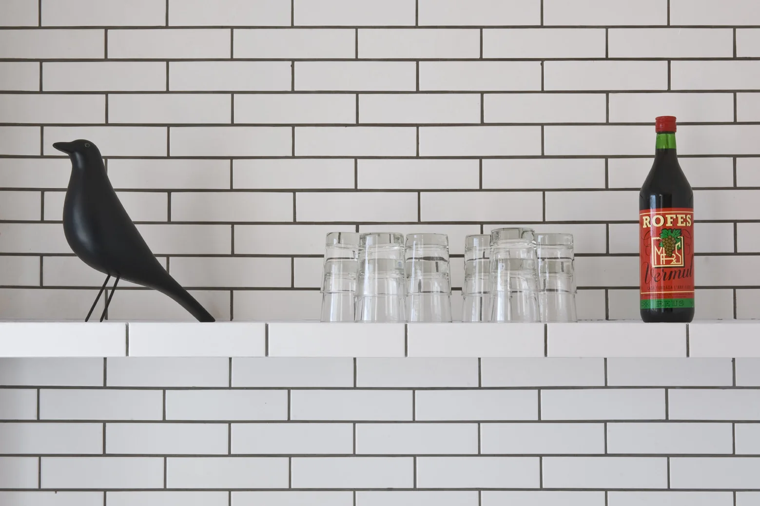 A white subway tiled wall and matching shelf styled with a black bird figurine, a stack of glass cups, and a vermouth bottle.