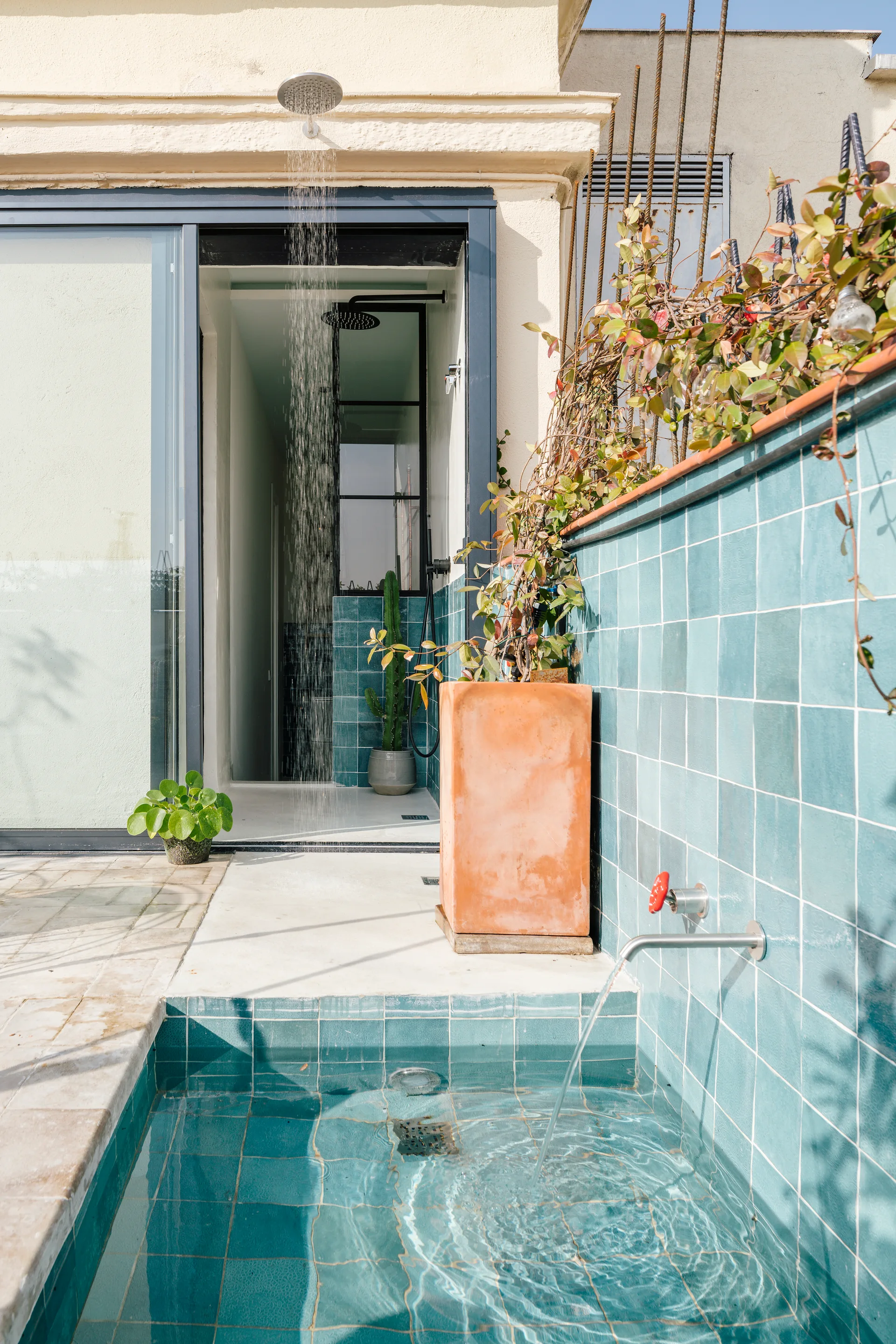 An outdoor patio featuring a small plunge pool and shower walled in glossy teal square tiles next to a terracotta planter.