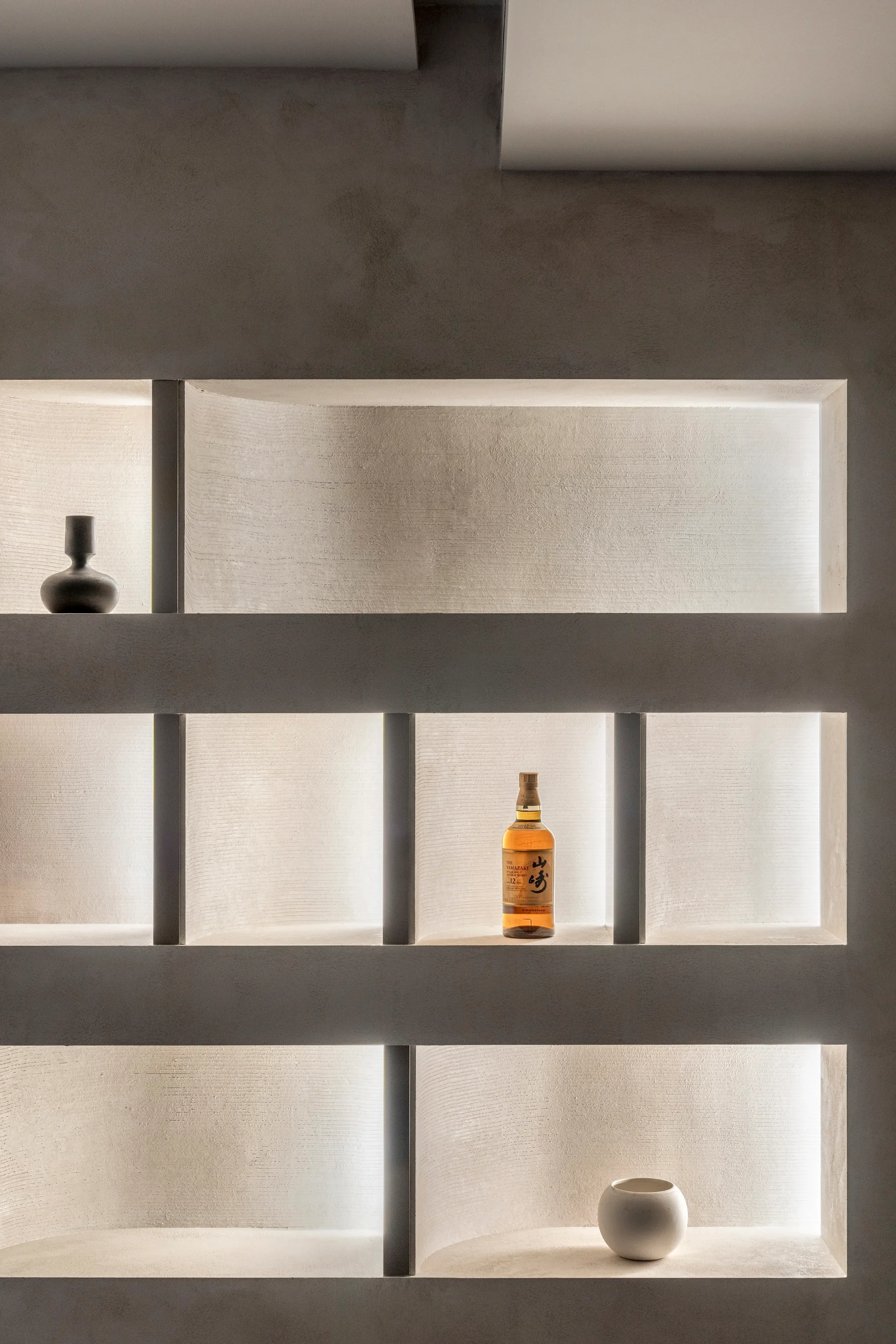 A minimalist beige plaster wall featuring built-in recessed shelves styled with a small dark vase, a bottle, and a white bowl under warm backlighting.