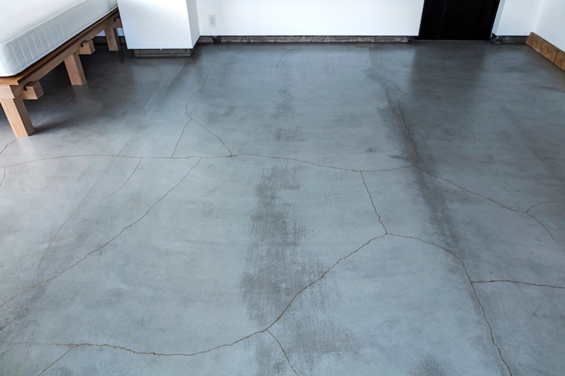 A minimalist room features a light grey concrete floor with natural cracks, a white wall, and a light wood bed frame.