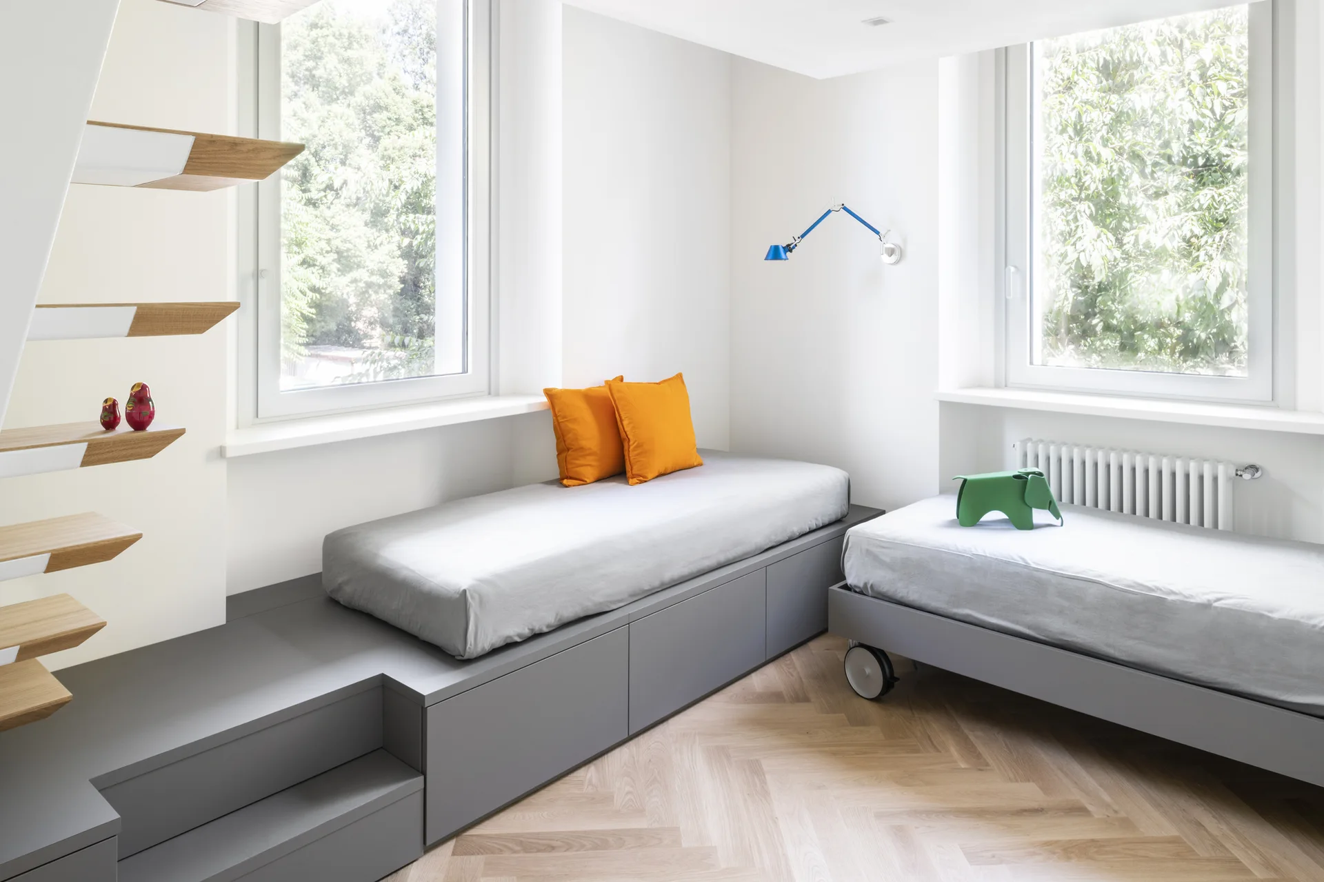 A bright kids' bedroom features a grey stepped storage platform bed, a secondary wheeled bed, and floating wooden stairs on herringbone flooring.