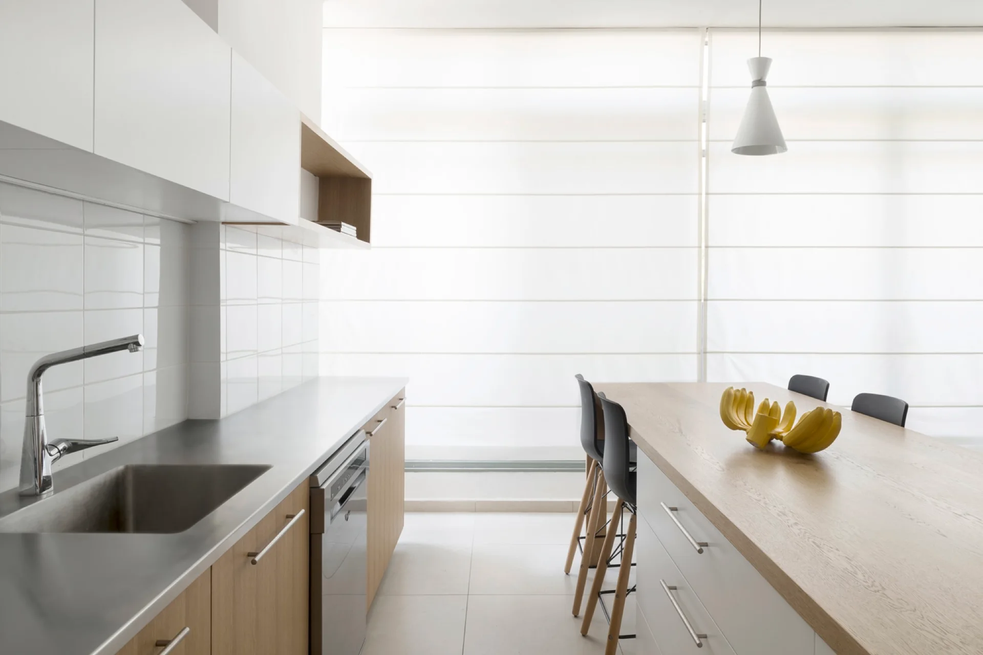 A minimalist kitchen features wood lower cabinets, white upper cabinets, a stainless steel sink countertop, and a matching wood-topped island with stools.