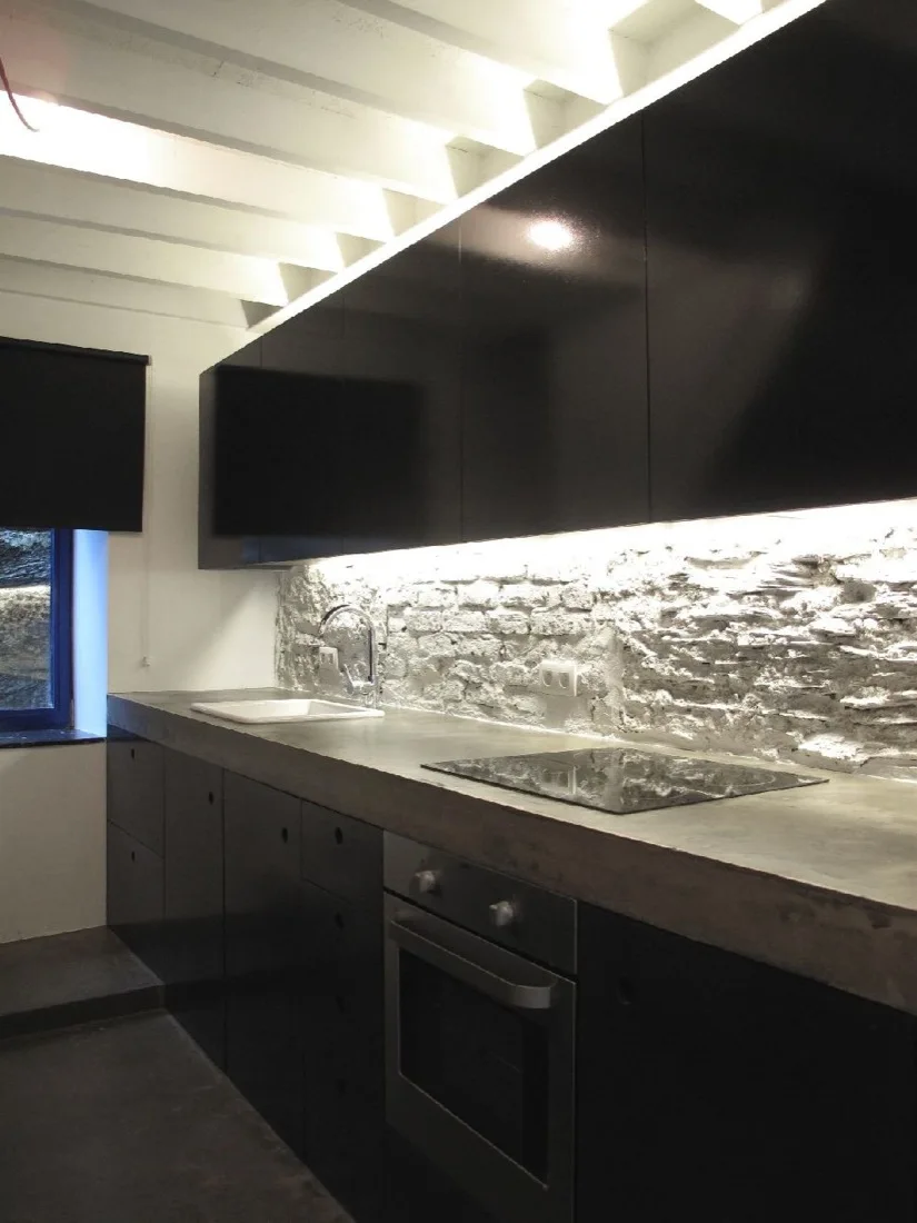 A modern kitchen features black flat-panel cabinetry, a thick concrete countertop, and a white textured stone backsplash illuminated by under-cabinet lighting.