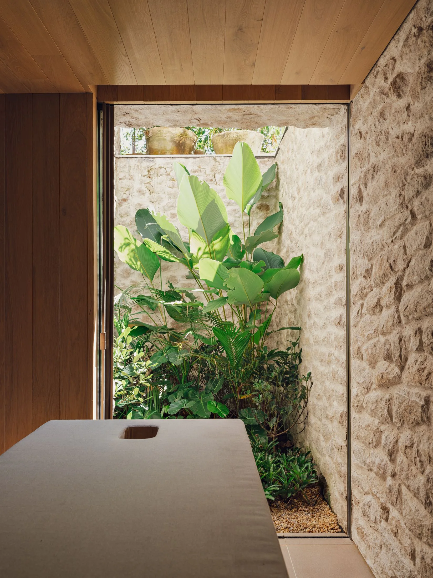 A spa treatment room features a beige massage table next to an open-air light well filled with green plants and bounded by a rough stone wall.
