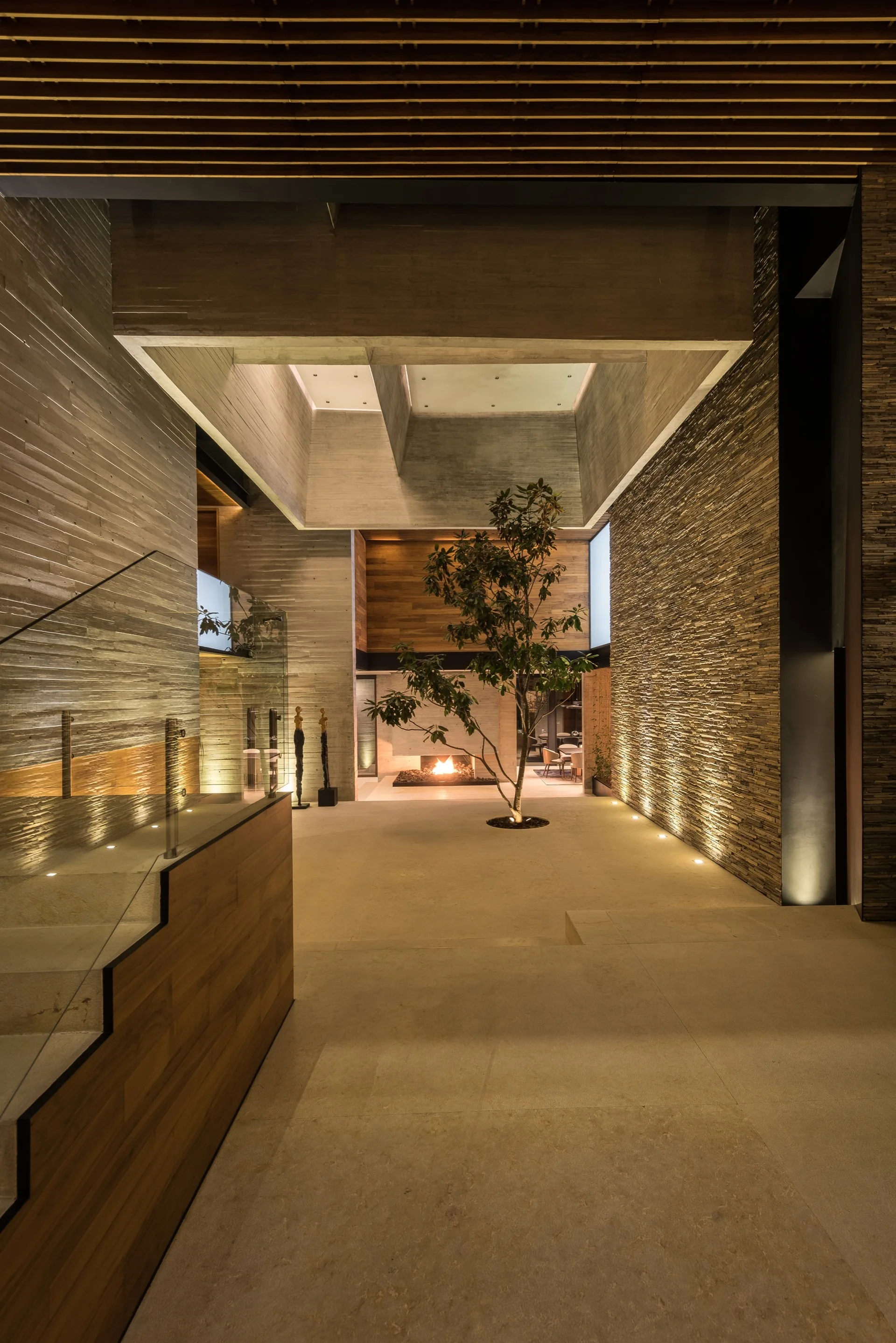 A modern foyer featuring a live green tree in the center, textured stone walls, warm uplighting, and a slatted wood ceiling.