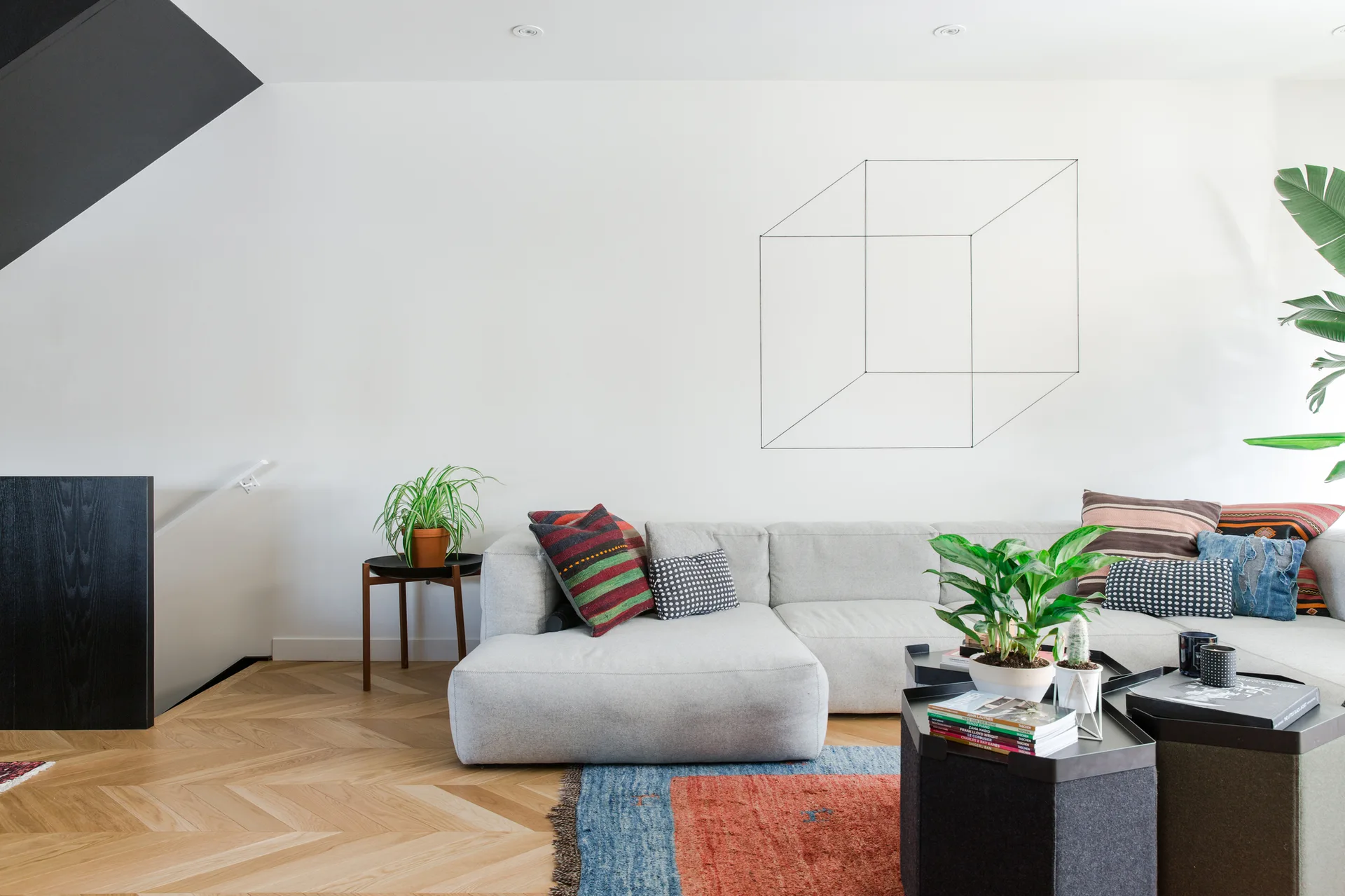 A light gray sectional sofa sits on herringbone wood flooring next to modular black coffee tables and a white wall featuring geometric line art.