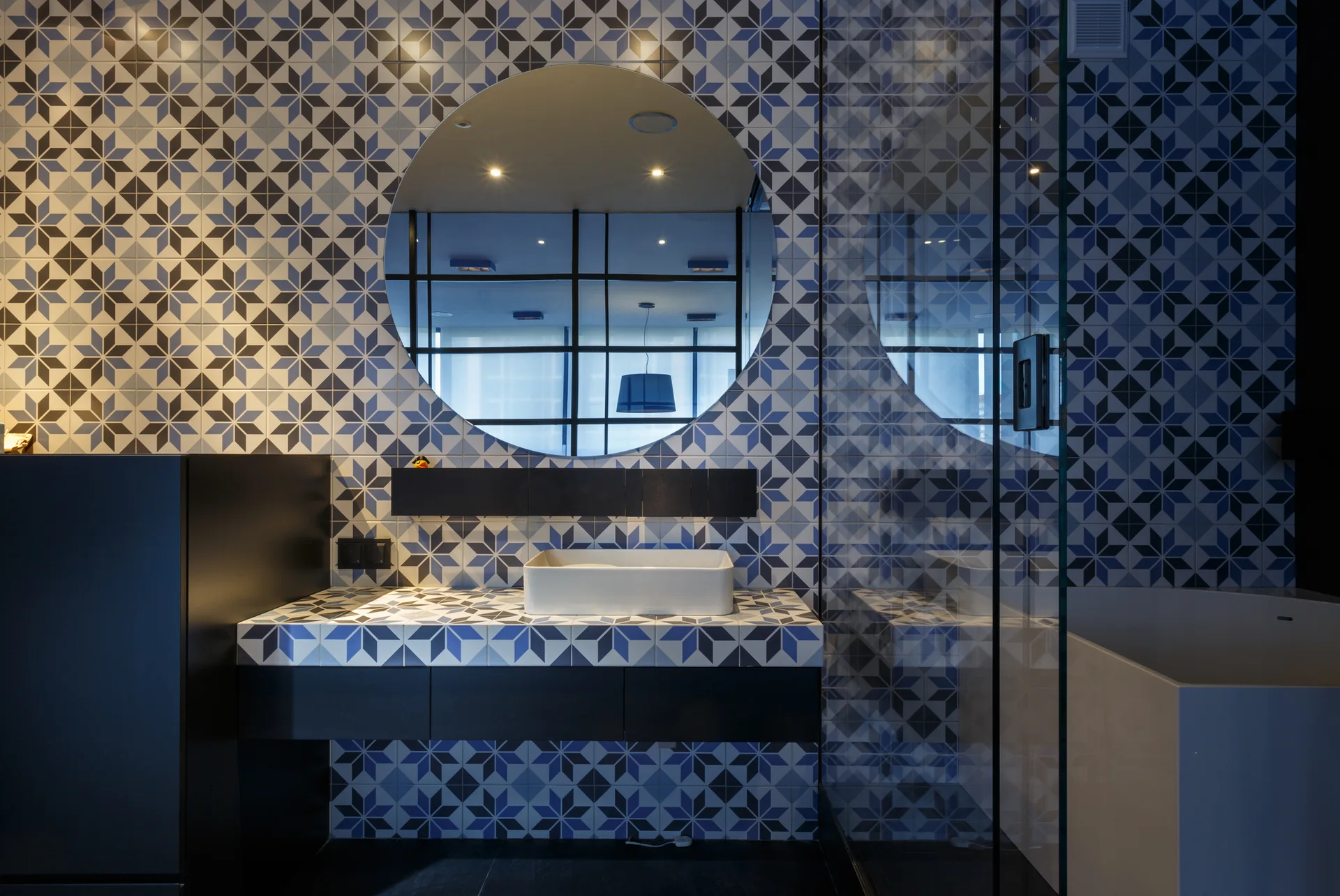 A modern bathroom features a vessel sink on a tiled vanity, geometric blue and white star patterned wall tiles, and a large round mirror.