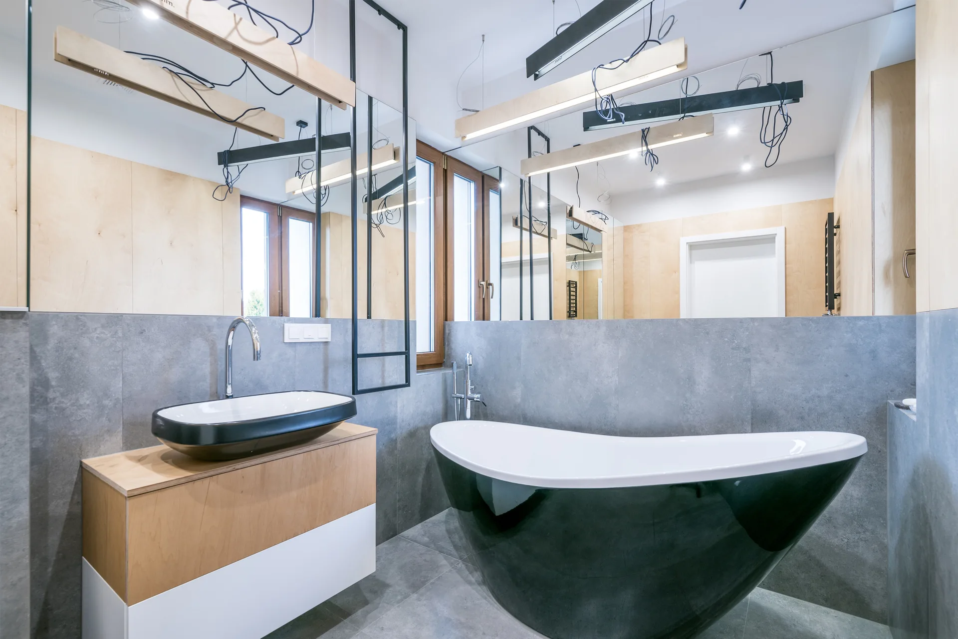 A modern bathroom featuring a black and white freestanding bathtub, a wood-faced floating vanity with a vessel sink, and large mirrors over grey stone tiles.