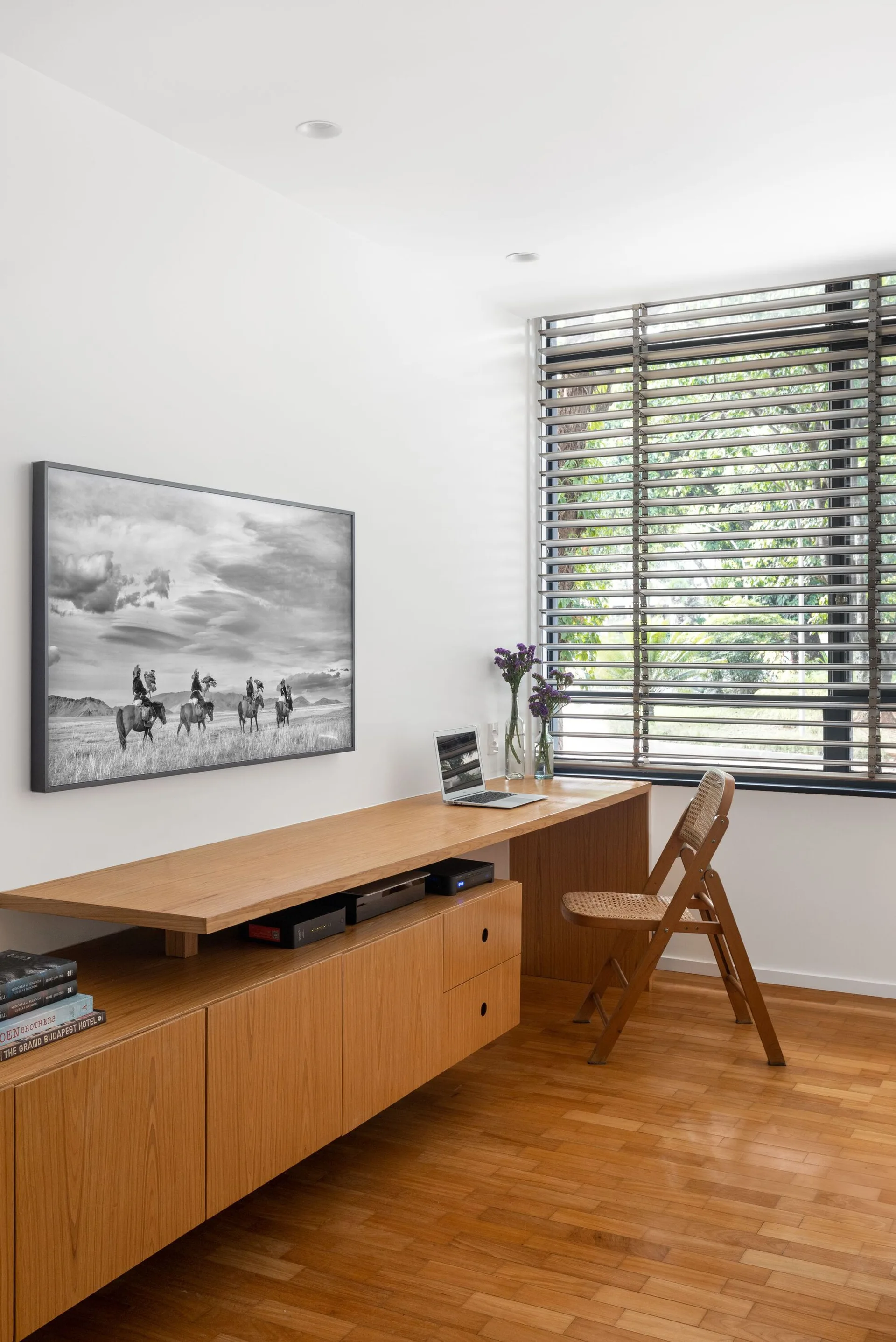 A minimalist home office features a long built-in wooden desk, a cane folding chair, and a large window with horizontal blinds.