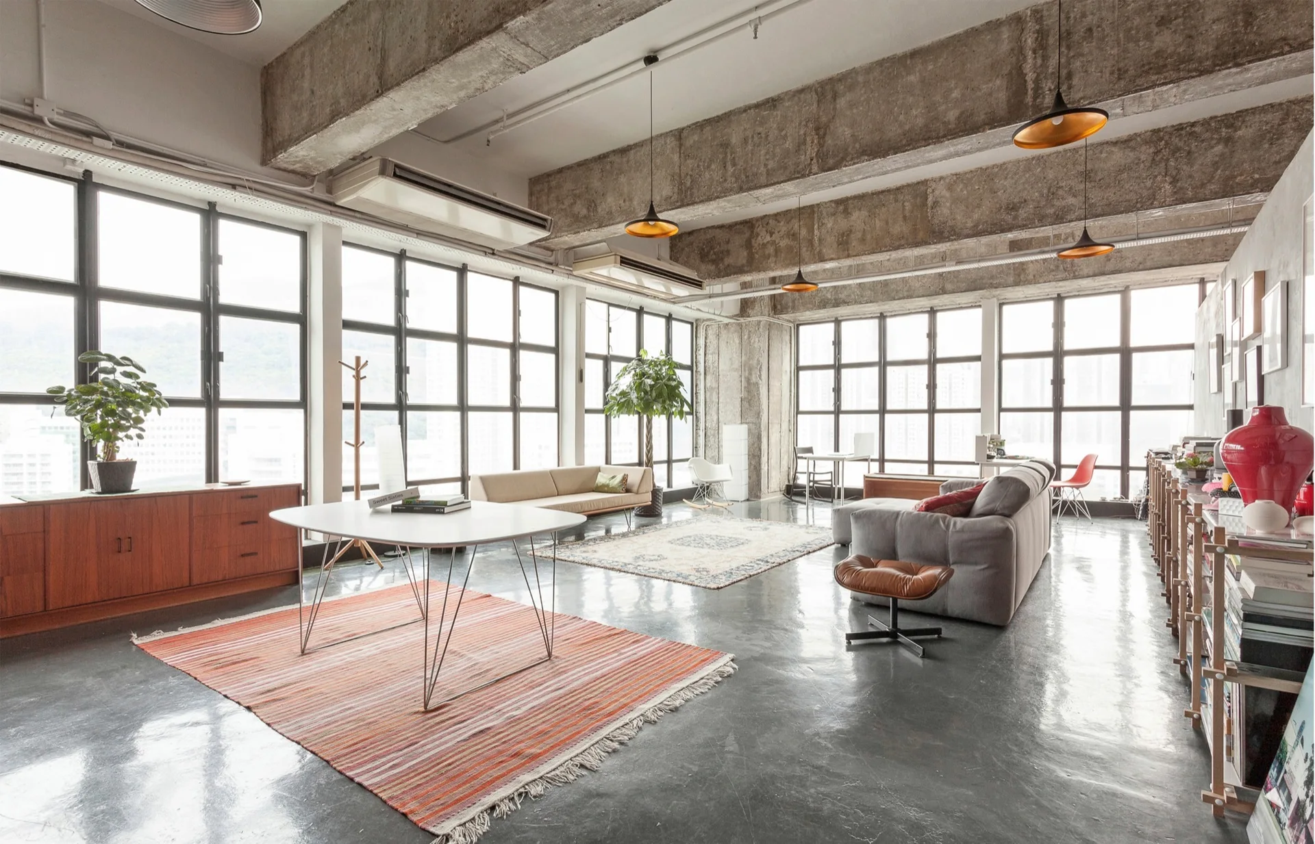 An open loft living space with exposed concrete ceiling beams, large metal grid windows, a grey sofa, and mid-century modern furniture on concrete floors.