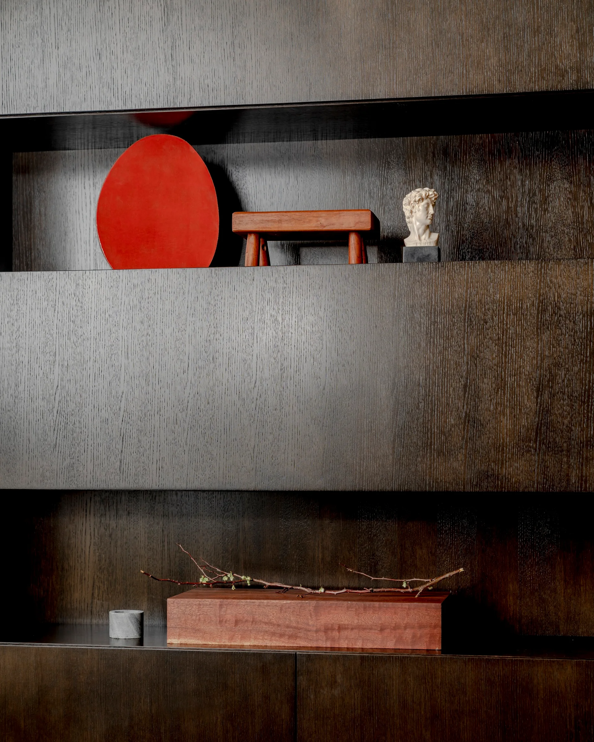 A close-up of dark brown wooden built-in shelves styled with a red circular plate, a small wooden bench, a miniature white bust, and a branch on a wooden block.