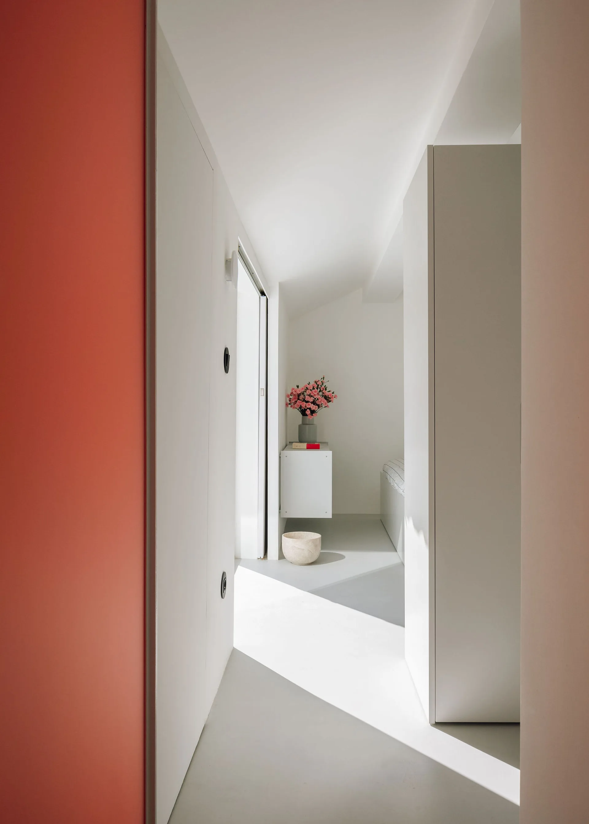 A minimalist white corridor features a bold coral accent wall, sunlight casting sharp shadows on the floor, and a floating cabinet with pink flowers.