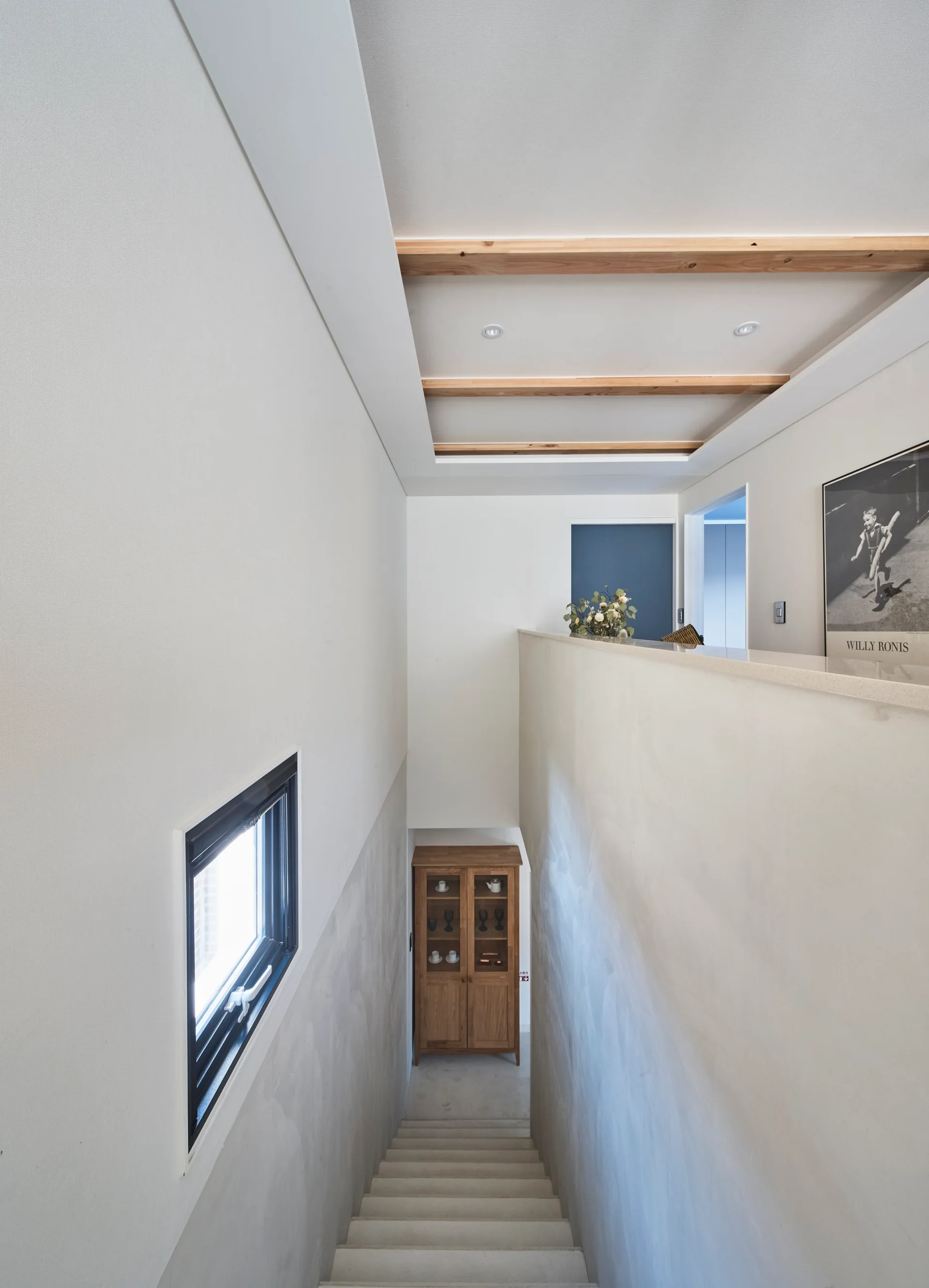 A minimalist staircase with textured plaster walls features a recessed ceiling with wood beams, a small window, and a wooden cabinet at the base.