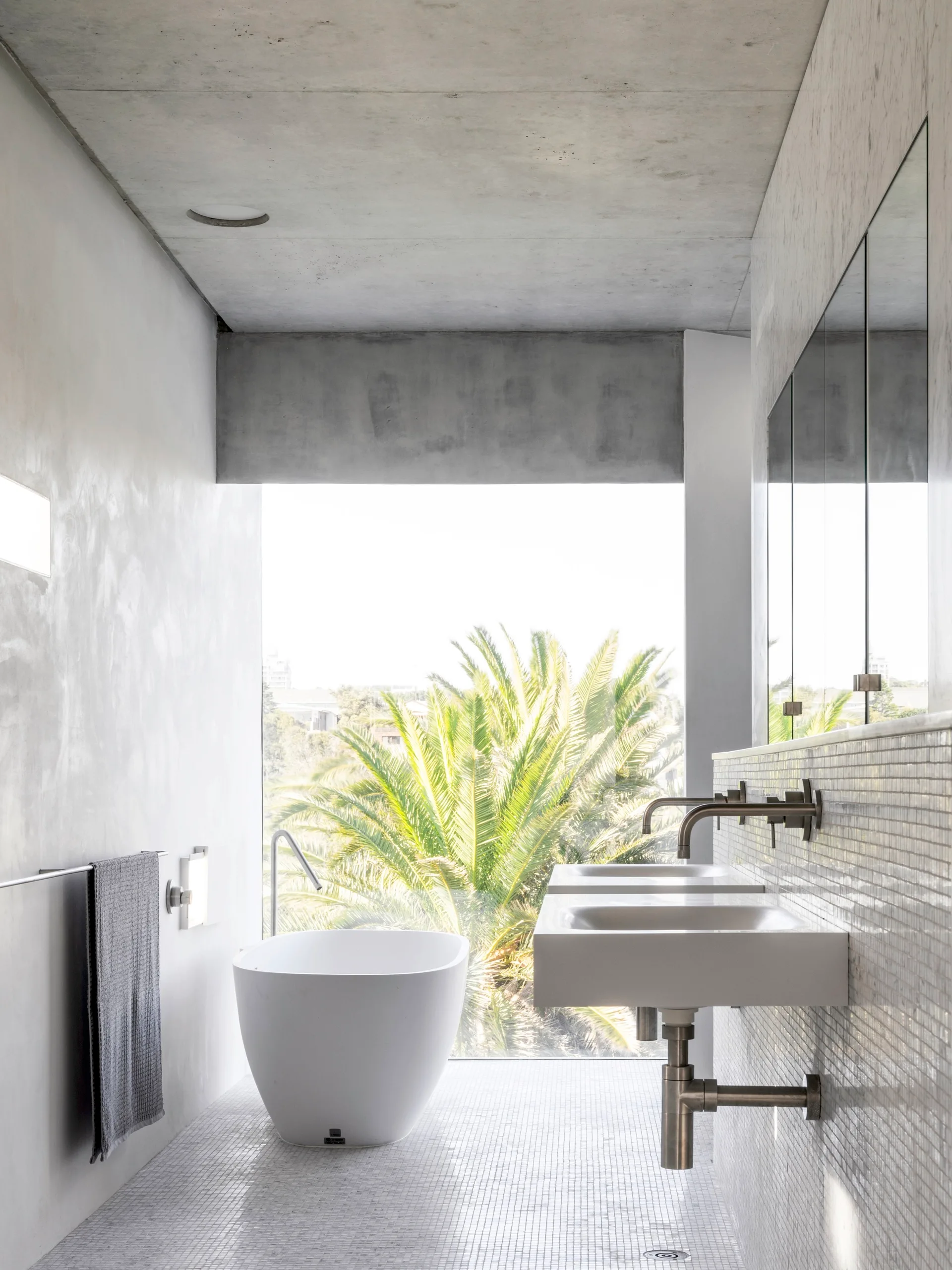 A minimalist bathroom featuring a white freestanding bathtub, concrete walls and ceiling, a wall-mounted vanity, and a large window looking out at a palm tree.