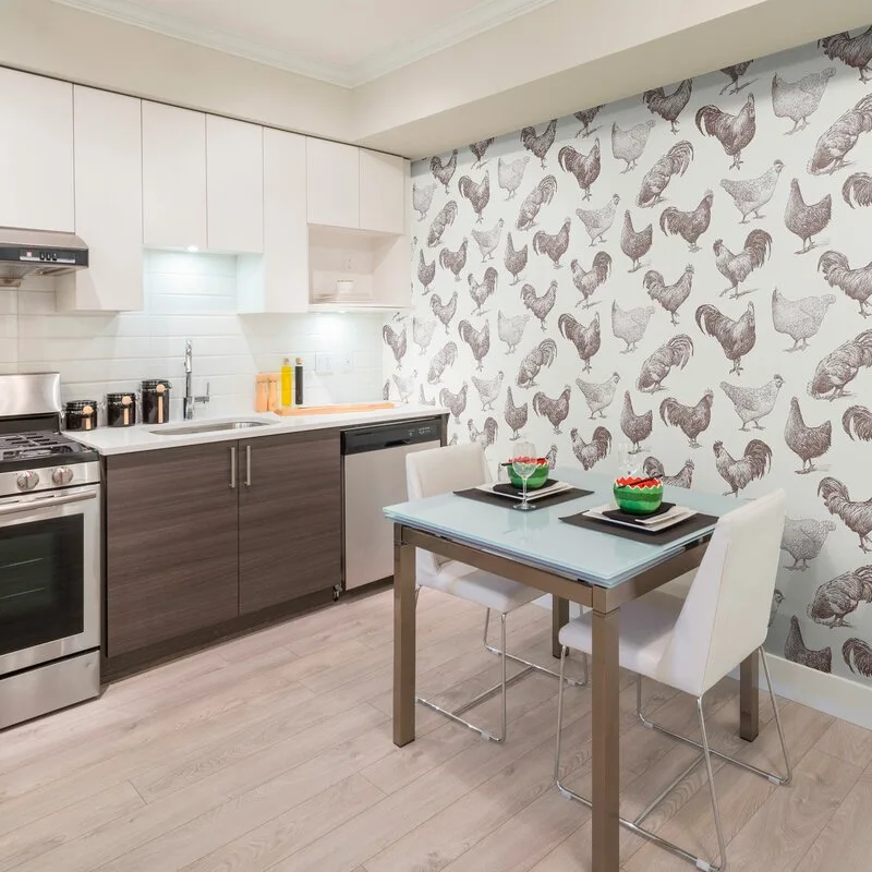 A small dining table with a glass top and white chairs sits next to a kitchen wall covered in black-and-white chicken pattern wallpaper.