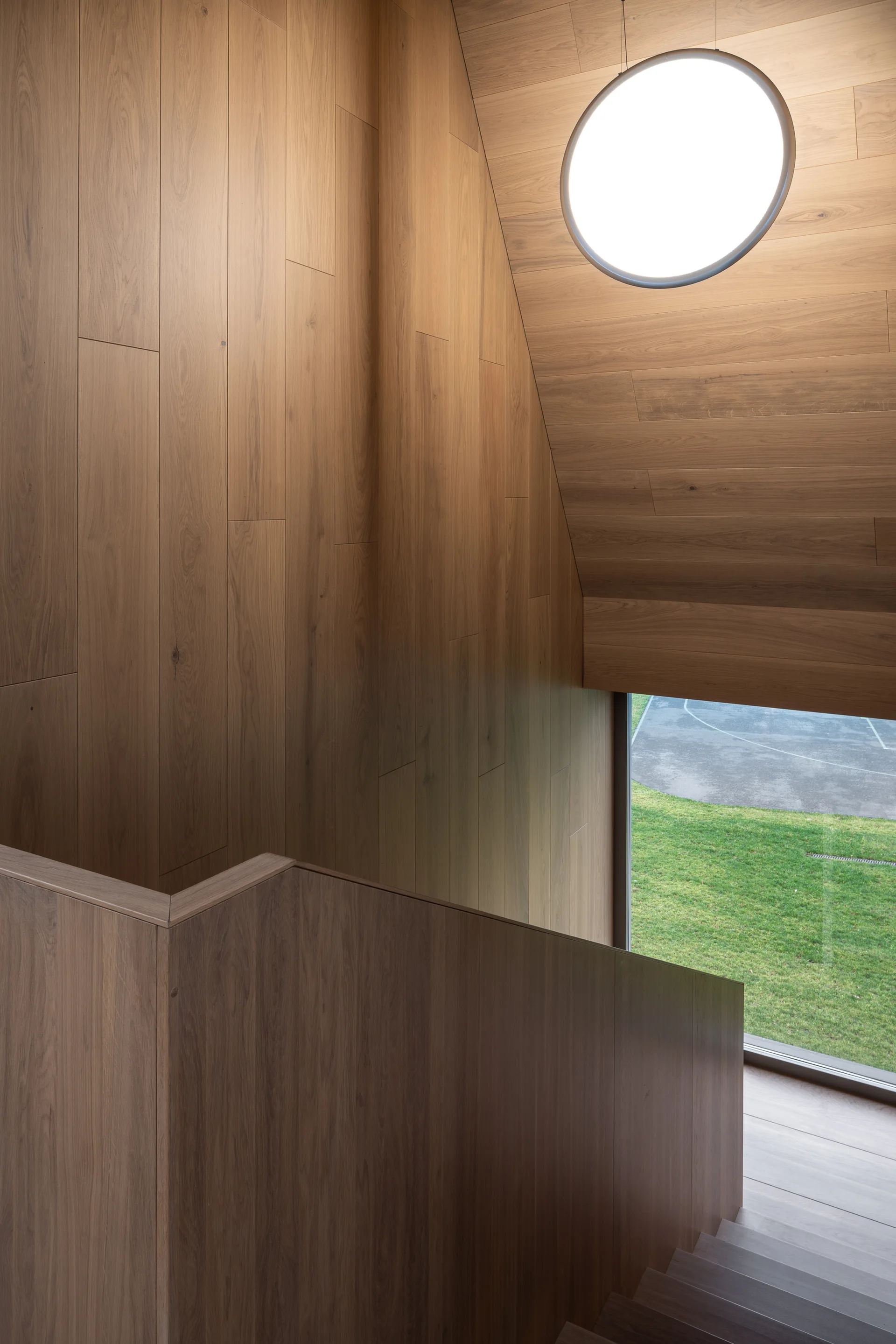 A modern minimalist staircase clad in warm wood paneling, featuring a sloped ceiling, a large glowing circular pendant light, and a window showing a lawn.