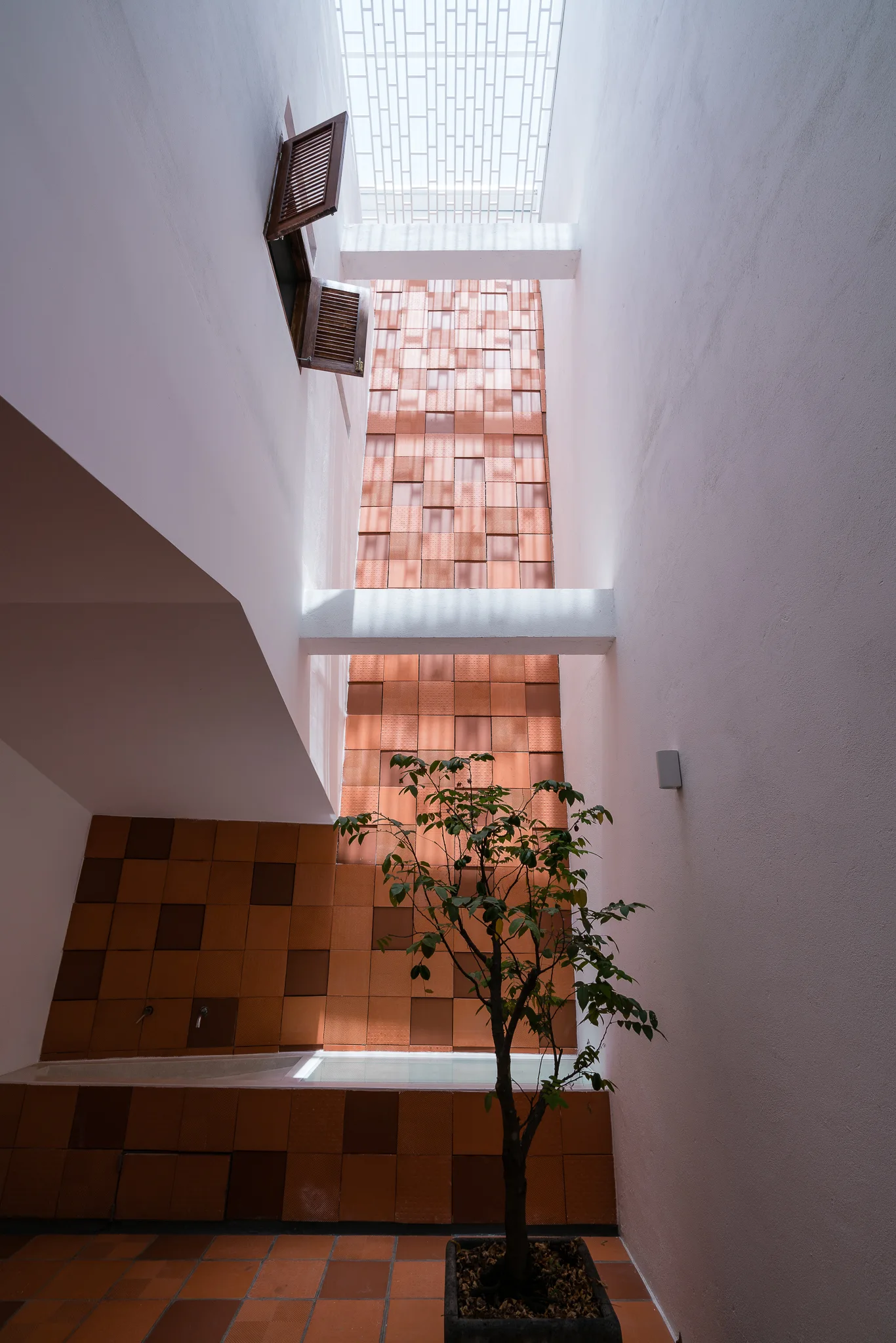 A tall indoor light well features a feature wall of square terracotta tiles, white plaster walls, an overhead skylight, and a potted green plant.