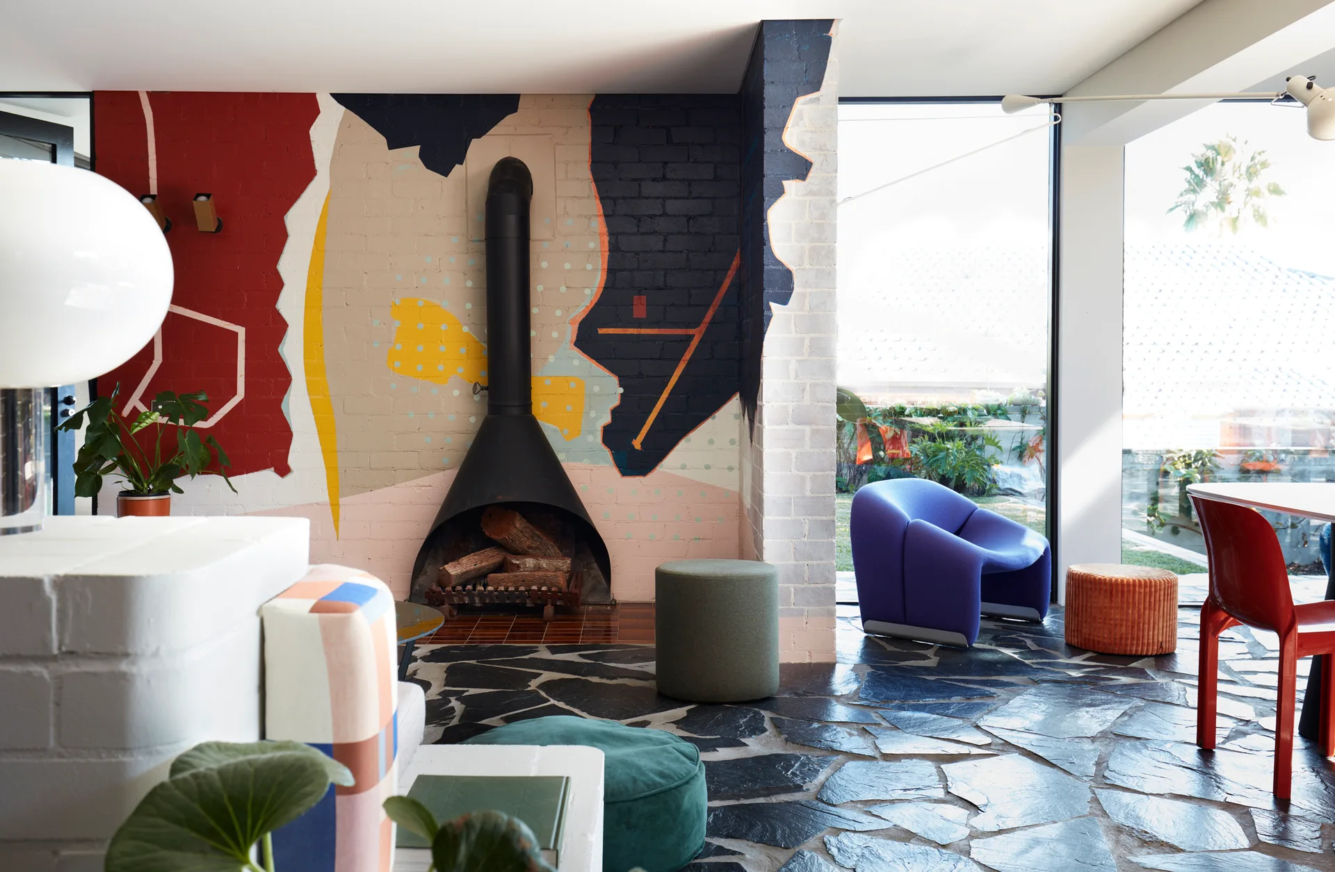 An eclectic living room featuring a conical black metal fireplace against an abstract painted brick wall, with dark flagstone flooring.
