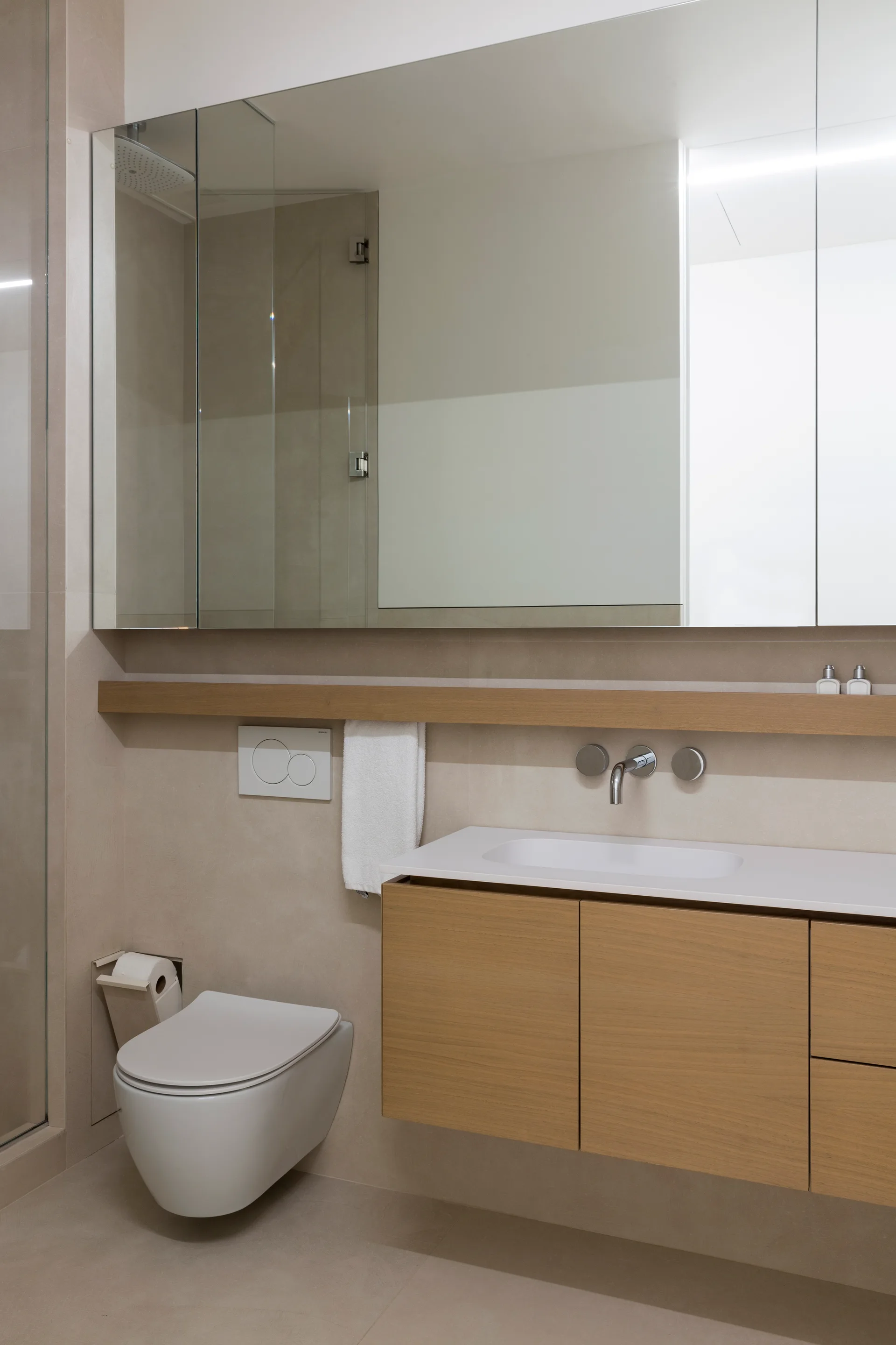 A contemporary bathroom featuring a floating light wood vanity, a wall-mounted toilet, and a large mirror against beige tile walls.