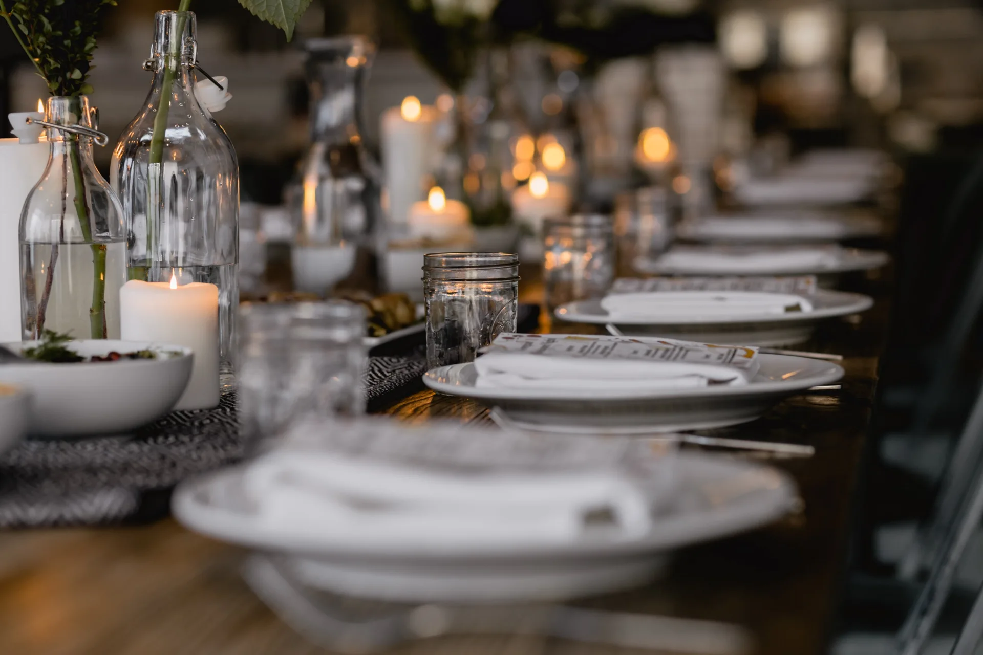 A long wooden dining table set with white ceramic plates, folded napkins, mason jars, glass bottles, and lit pillar candles.