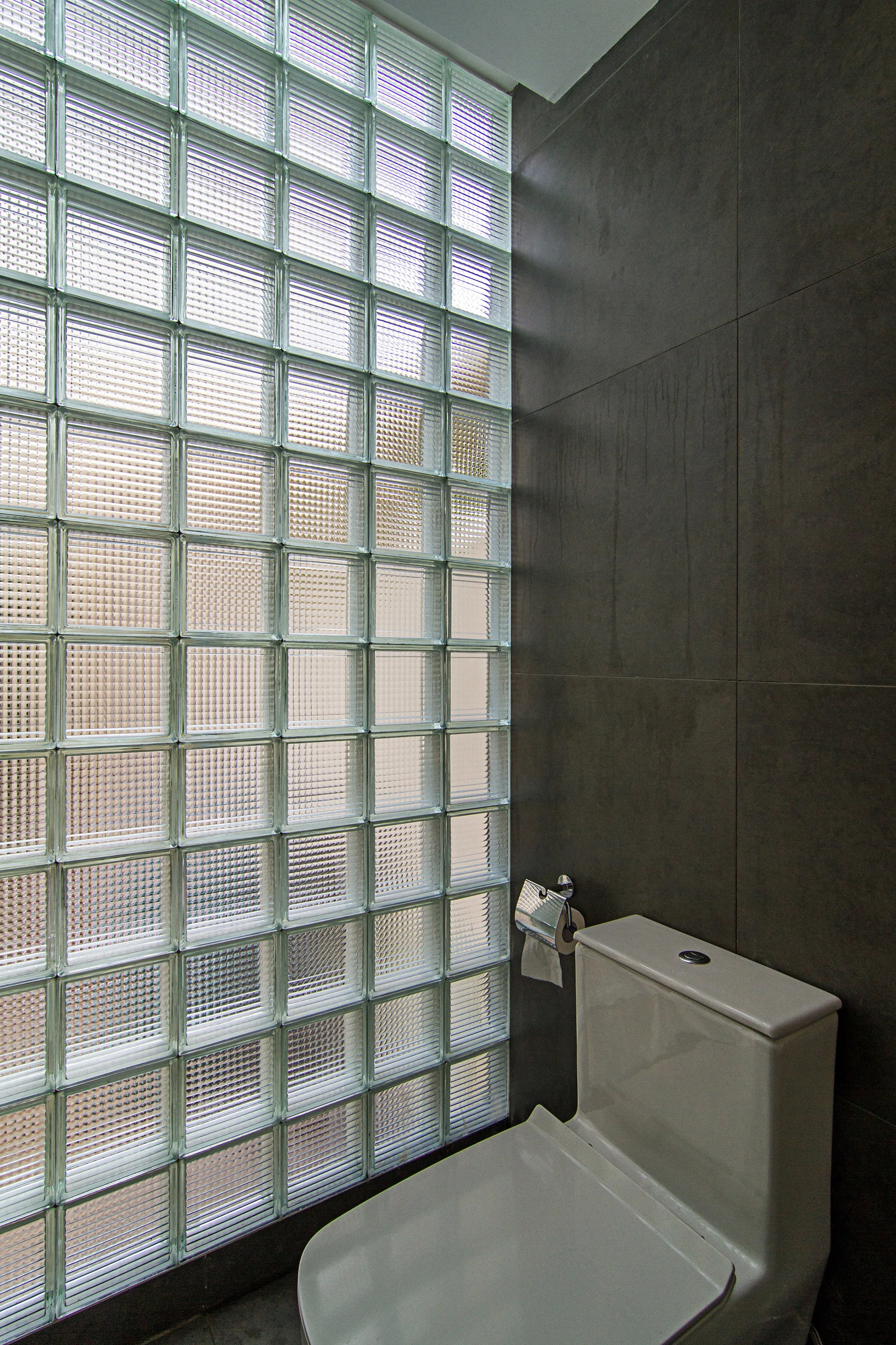 A modern bathroom featuring a white toilet set against a dark grey tiled wall next to a large translucent glass block window.