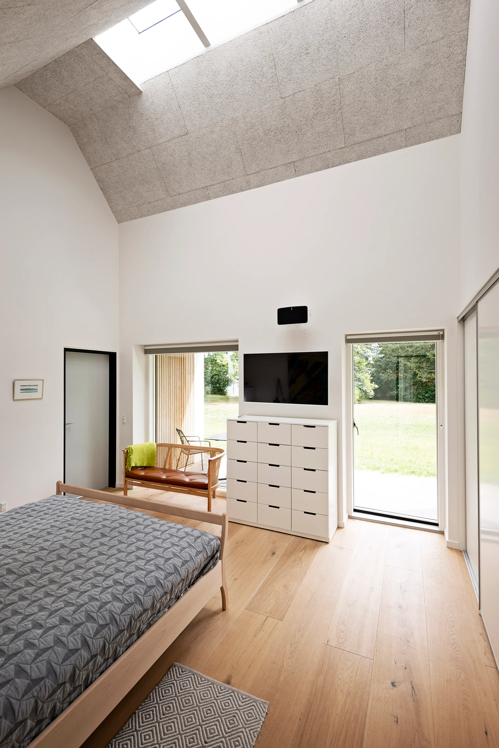 A minimalist bedroom featuring a vaulted ceiling with a skylight, a wooden bed, a white dresser, and glass doors opening to a yard.
