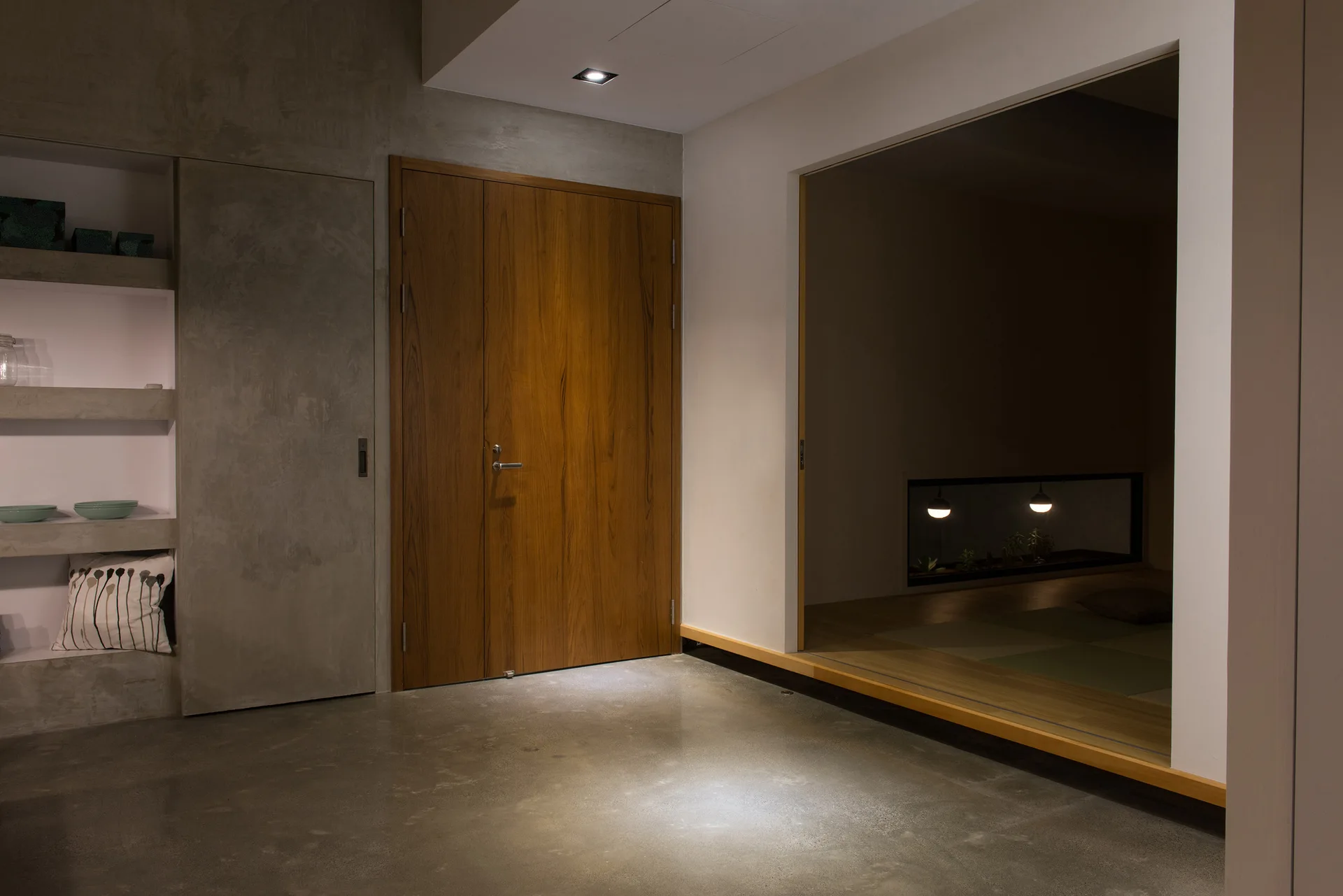 A minimalist entryway with a polished concrete floor, concrete walls featuring recessed shelves, a wood door, and an open doorway to a raised room.