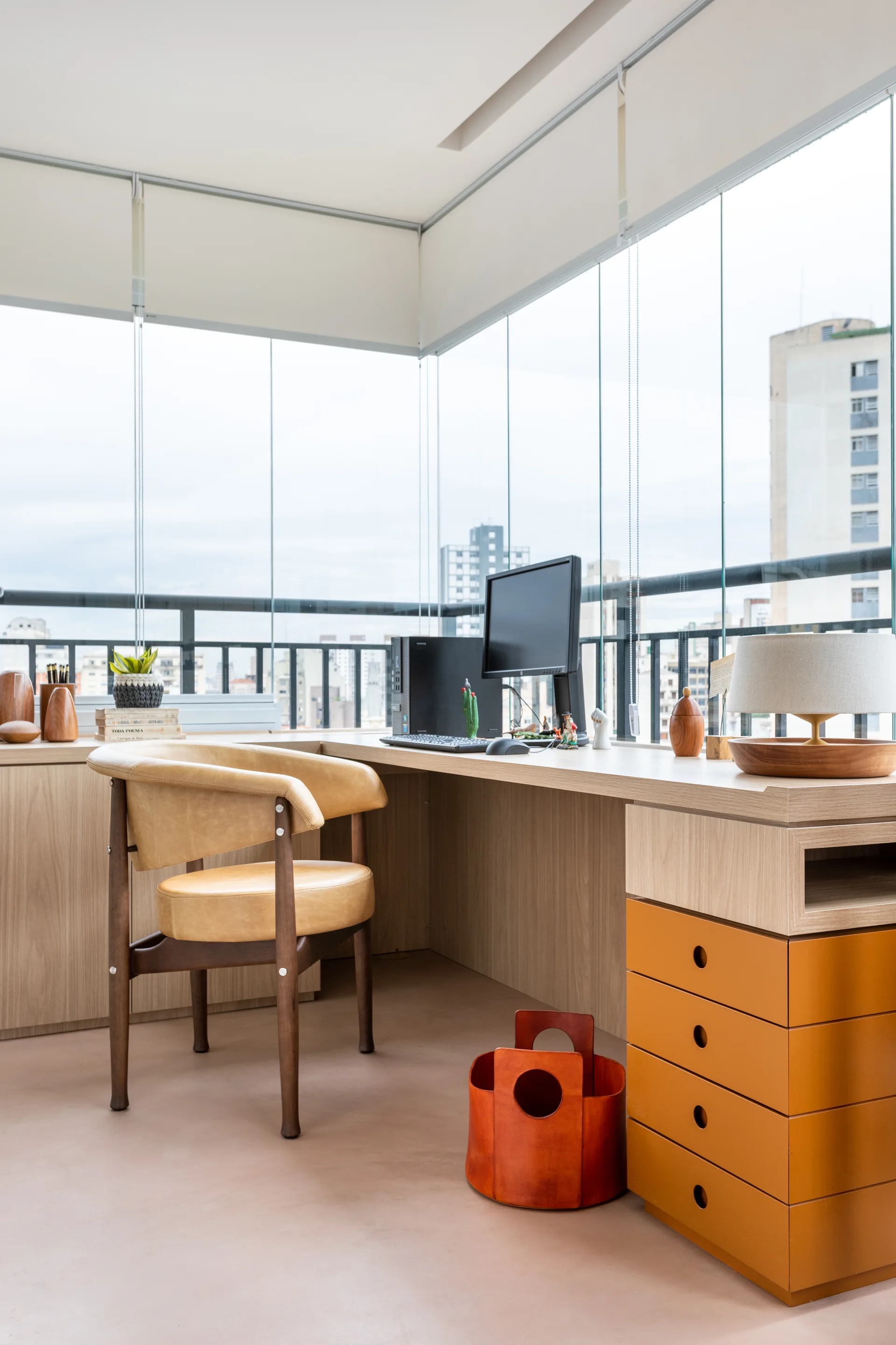 A modern home office on a glass-enclosed balcony features a light wood corner desk, a tan leather chair, and a stack of orange drawers.